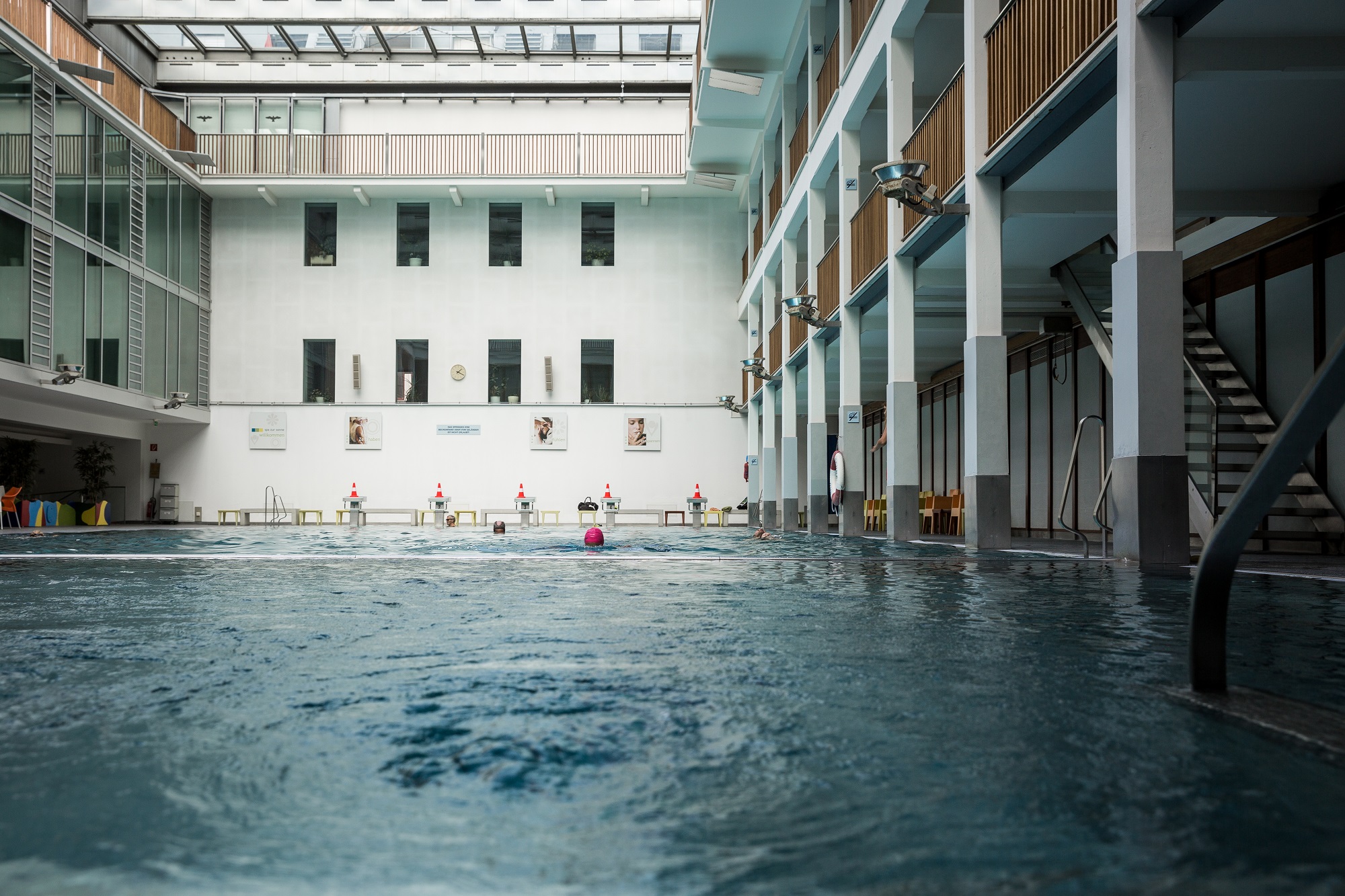 An indoor swimming pool with a person swimming laps. The pool is surrounded by a white-walled building with a glass roof.