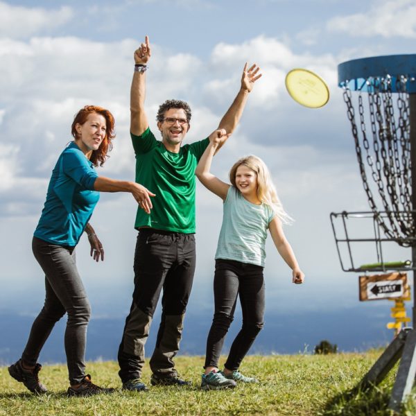 A family of three plays disc golf on the Schöckl plateau, with the father throwing a disc towards a basket labeled "START".