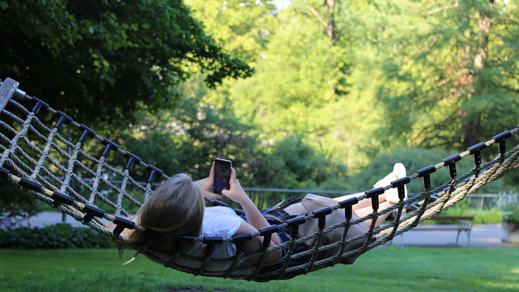 A woman lies in a hammock, looking at her phone. The hammock is in a grassy area with trees in the background.