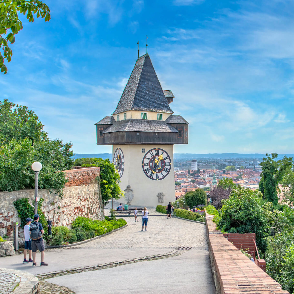 Sommerlicher Blick vom Schloßberg über Uhrturm auf Graz. Mehrere Touristen halten sich vor Uhrturm auf.