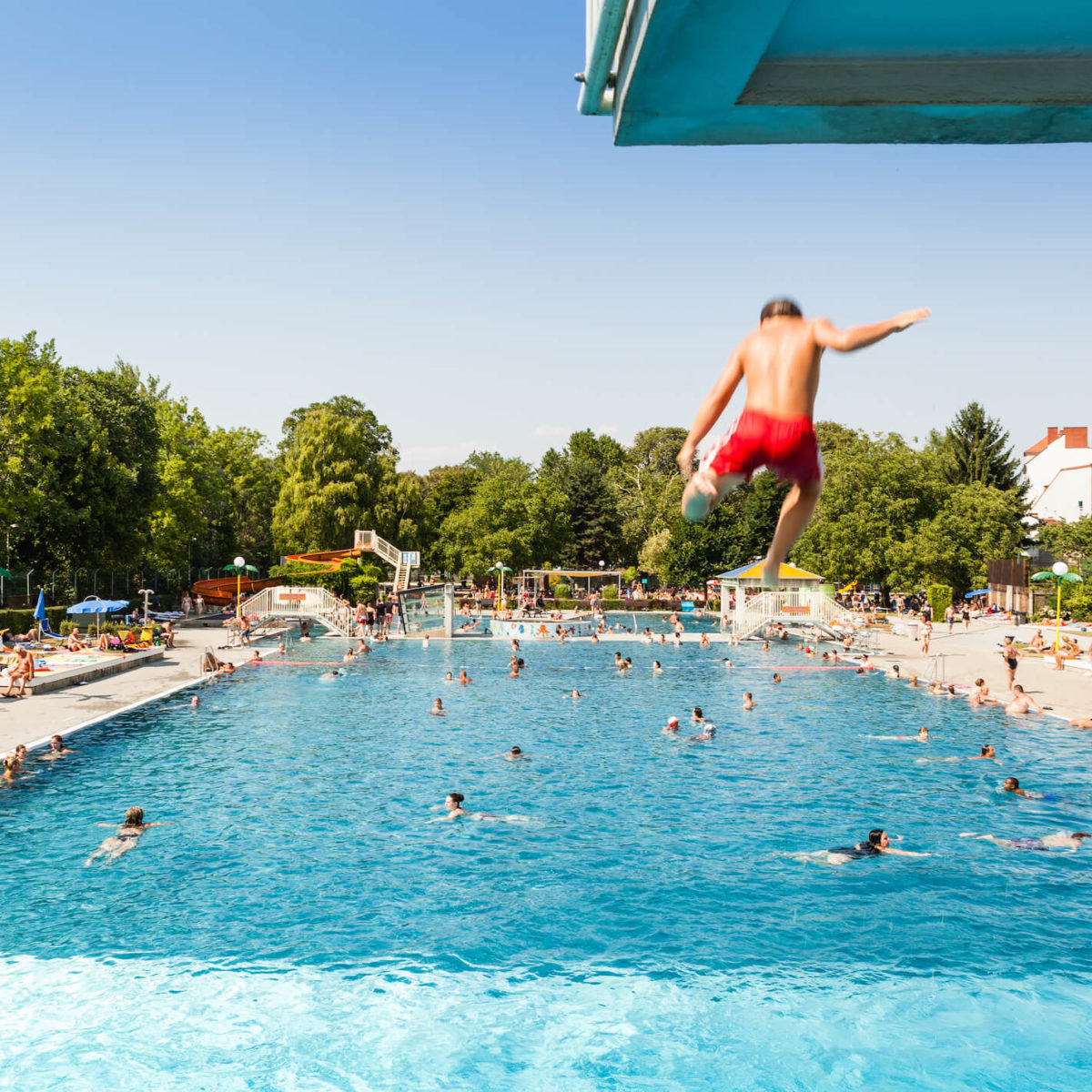 A young man jumps from a diving platform into a crowded outdoor swimming pool at Augartenbad.