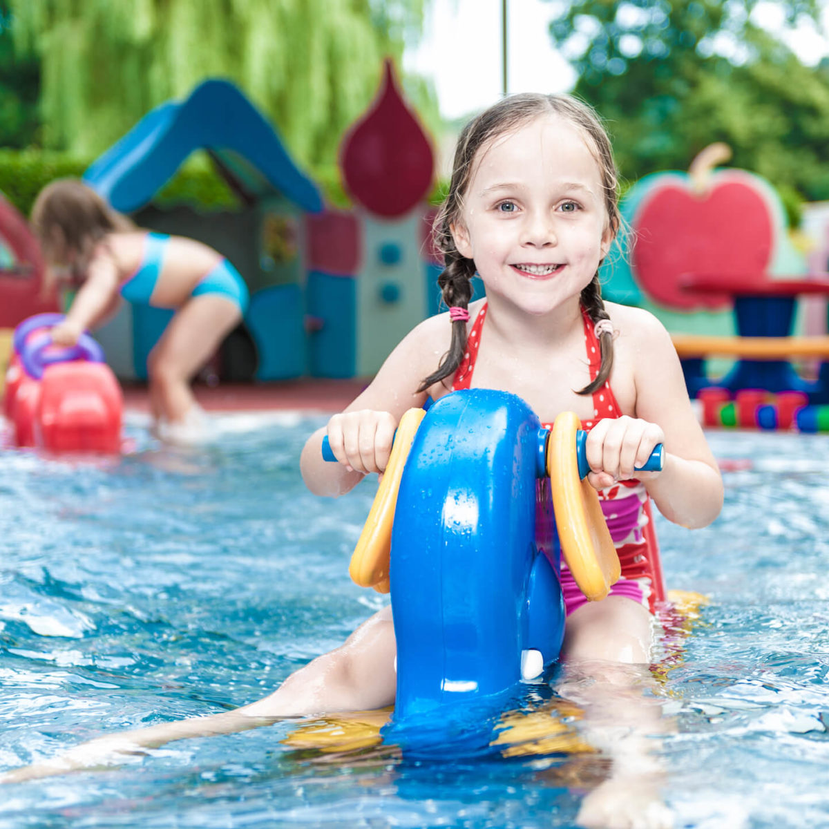 A young girl smiles as she rides a blue toy in a shallow pool. Another girl is visible in the background.