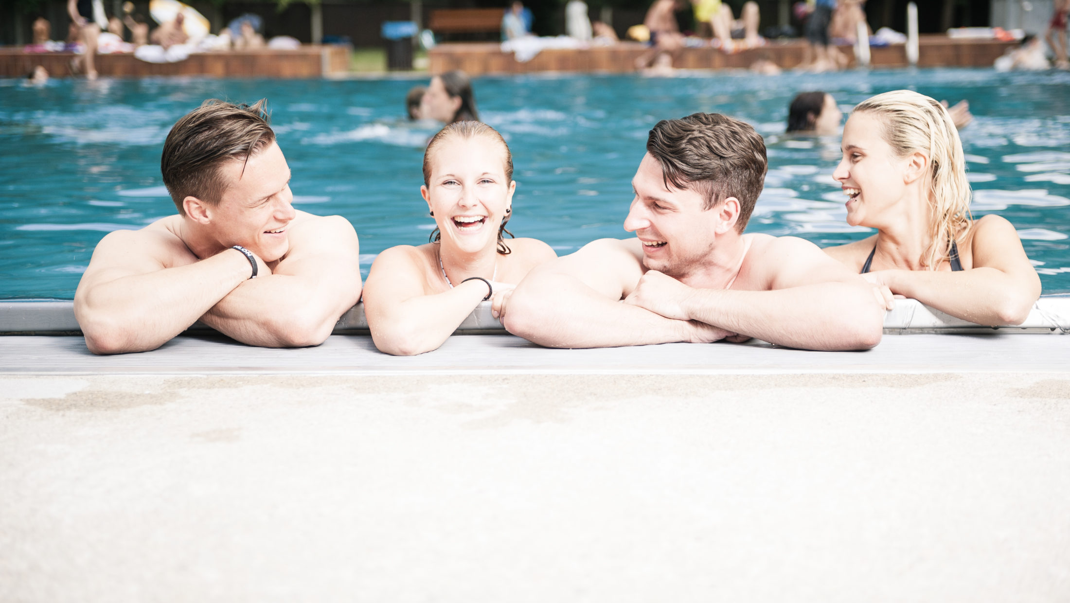 Four young adults laugh and chat while leaning on the edge of a swimming pool at the Margaretenbad.