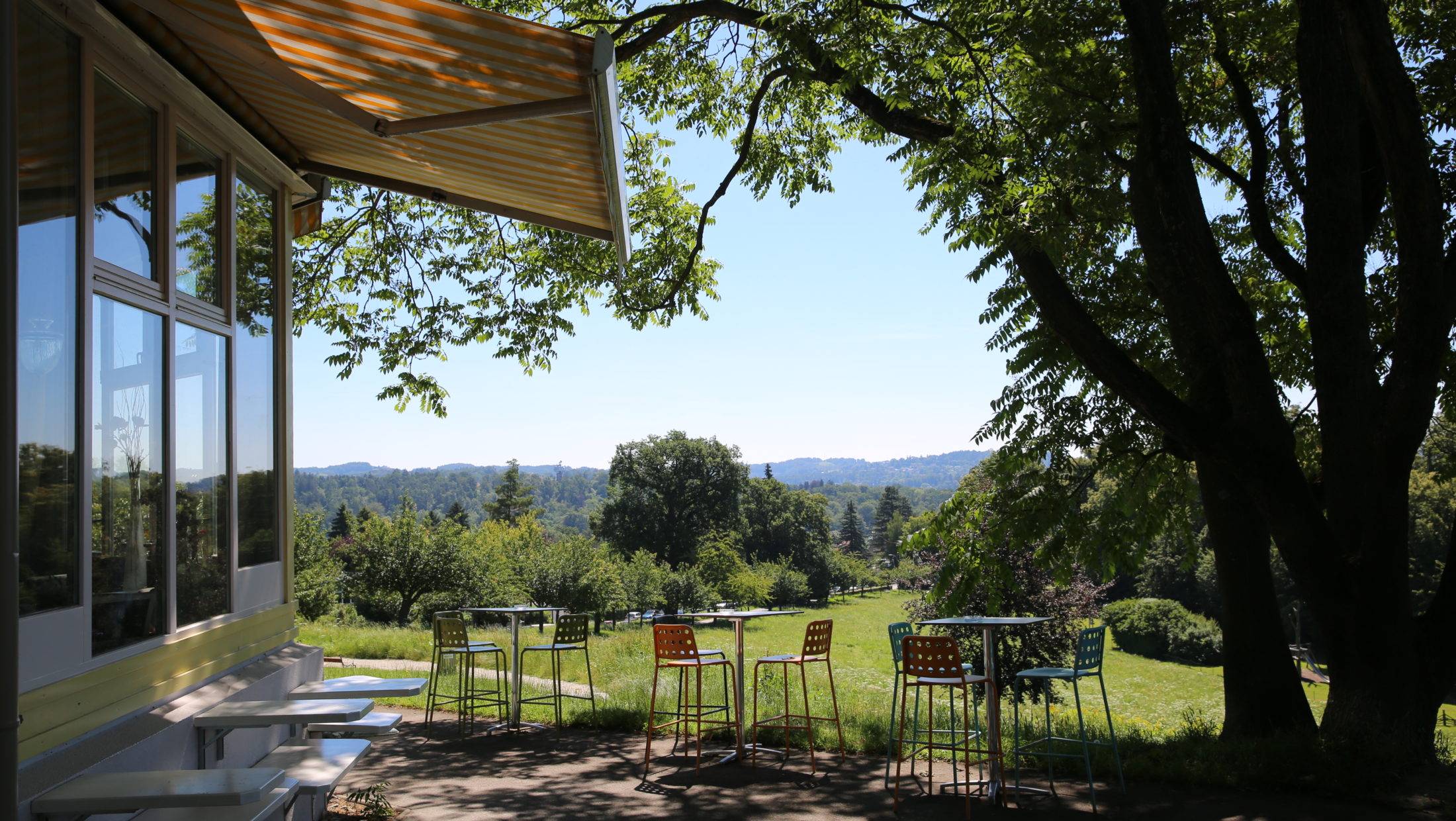 Empty patio with tables and chairs at Café Rosenhain, overlooking a grassy field and distant hills.