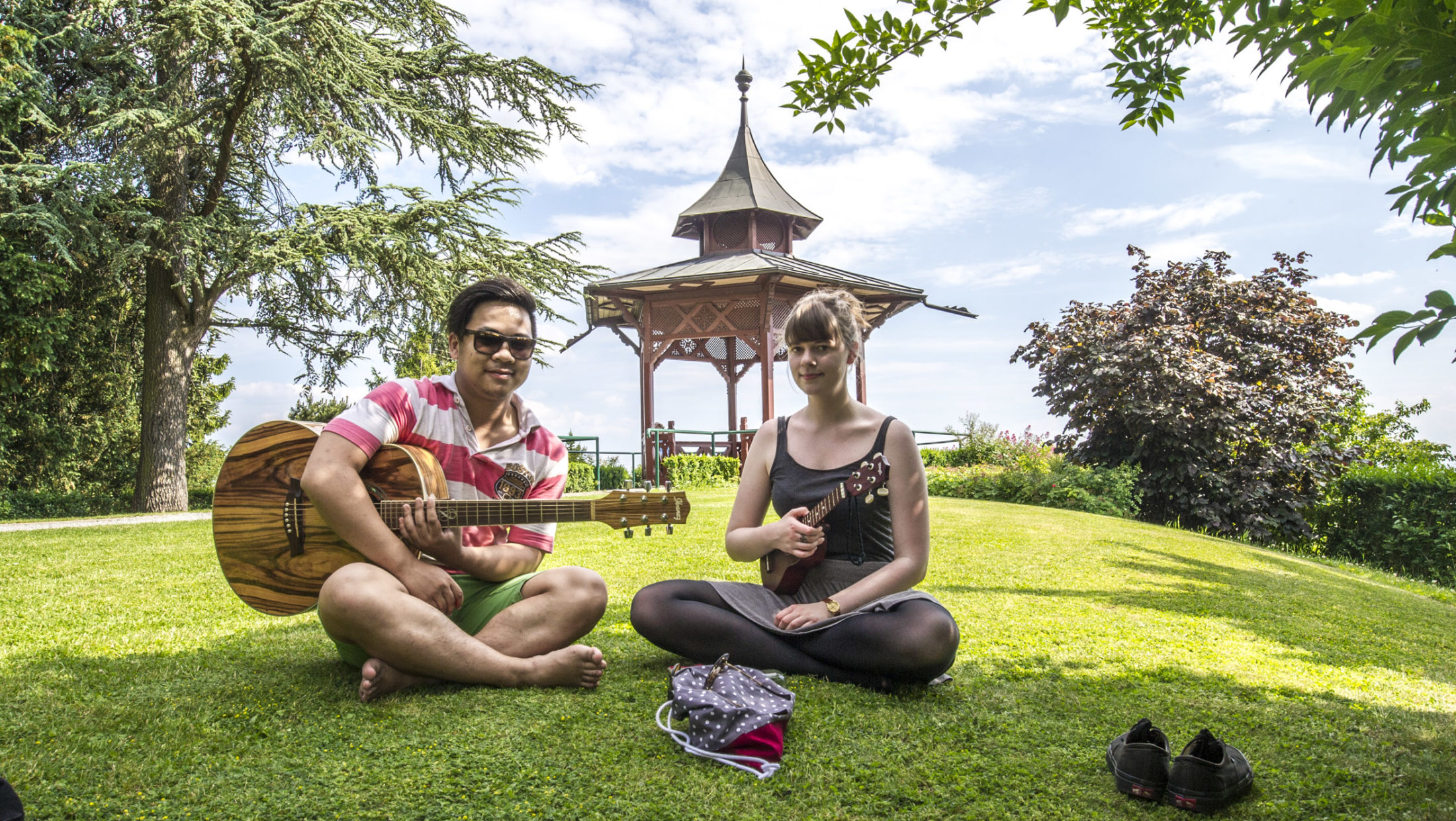 Junges Paar mit Gitarre und Ukulele sitzt in der Wiese vor chinesischem Pavillon am Schloßberg