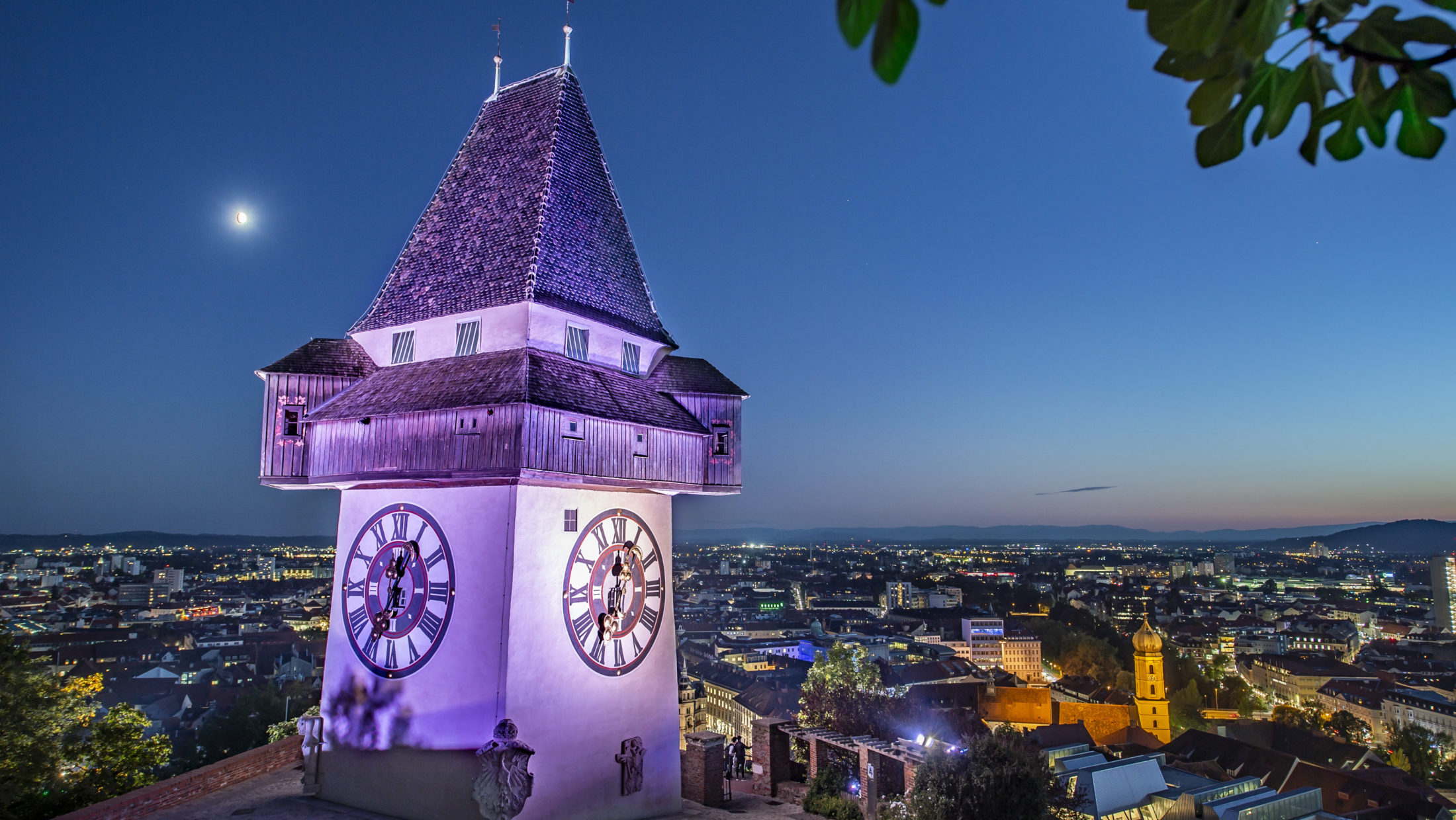 Violett beleuchteter Uhrturm am in der Nacht des Weltmädchentages, im Hintergrund nächtliches Graz.