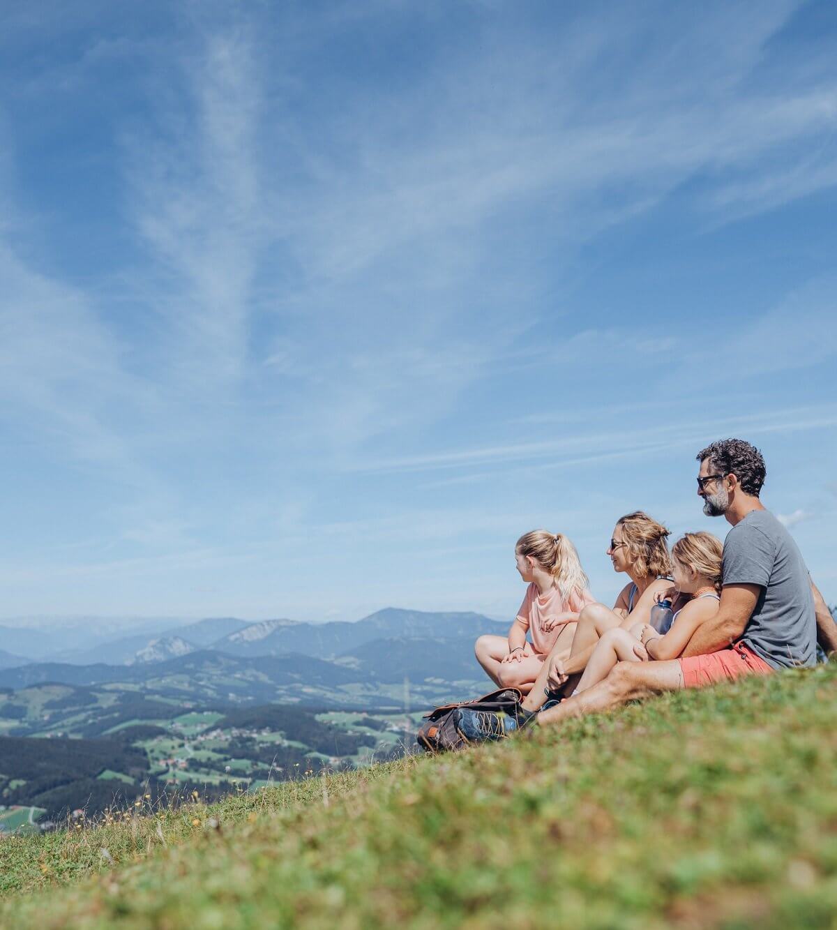 Familie mit zwei Mädchen sitzt am Gipfel des Schöckls in einer Wiese und blickt ins Tal