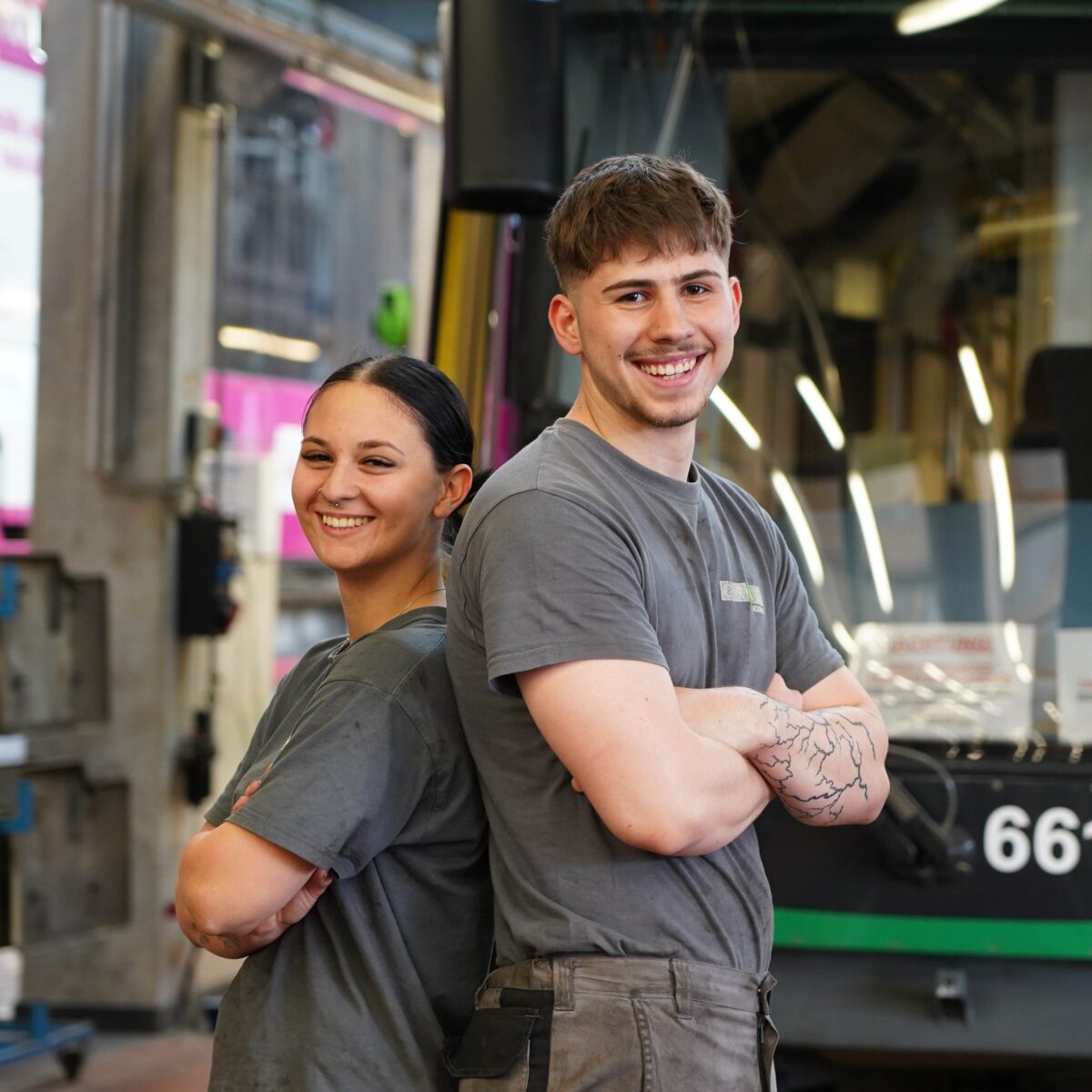 Two smiling mechanics stand back to back in a workshop. A tram is visible in the background.