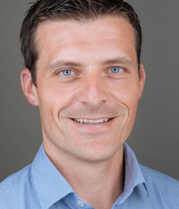 Headshot of Thomas Kalcher, a man with short brown hair, wearing a blue dress shirt and smiling.