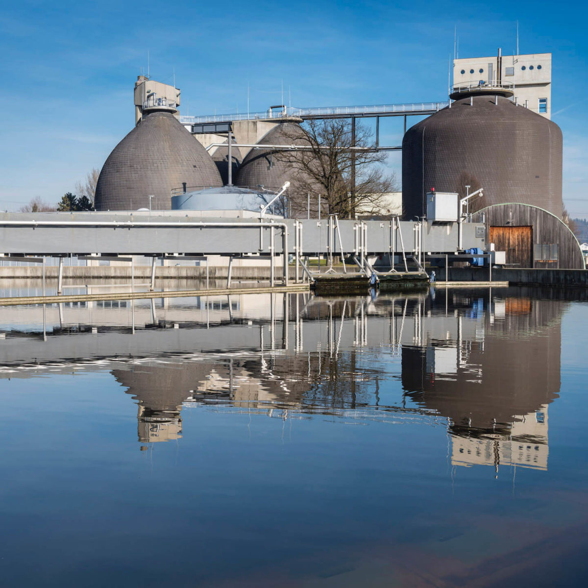 Sewage treatment plant with three large, domed tanks reflected in a still body of water.