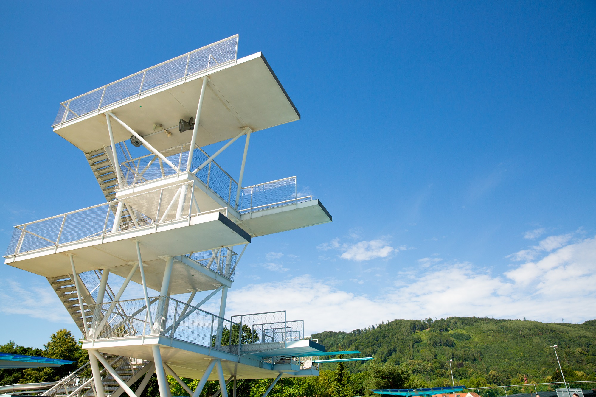 A white diving platform with multiple levels and a ladder, seen from below against a blue sky with white clouds.