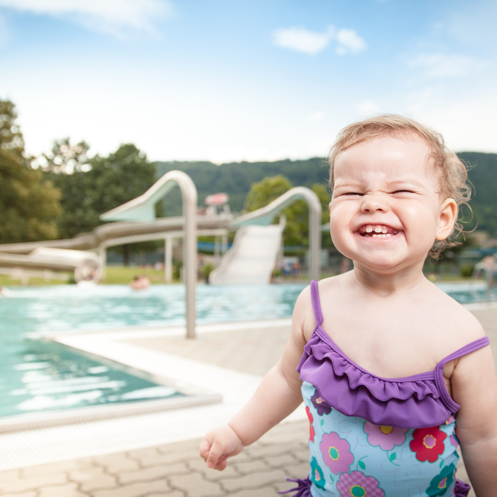 Kleines lachendes Mädchen in farbenfrohem Badeanzug lächelt in Auster Freibad fröhlich in Kamera.