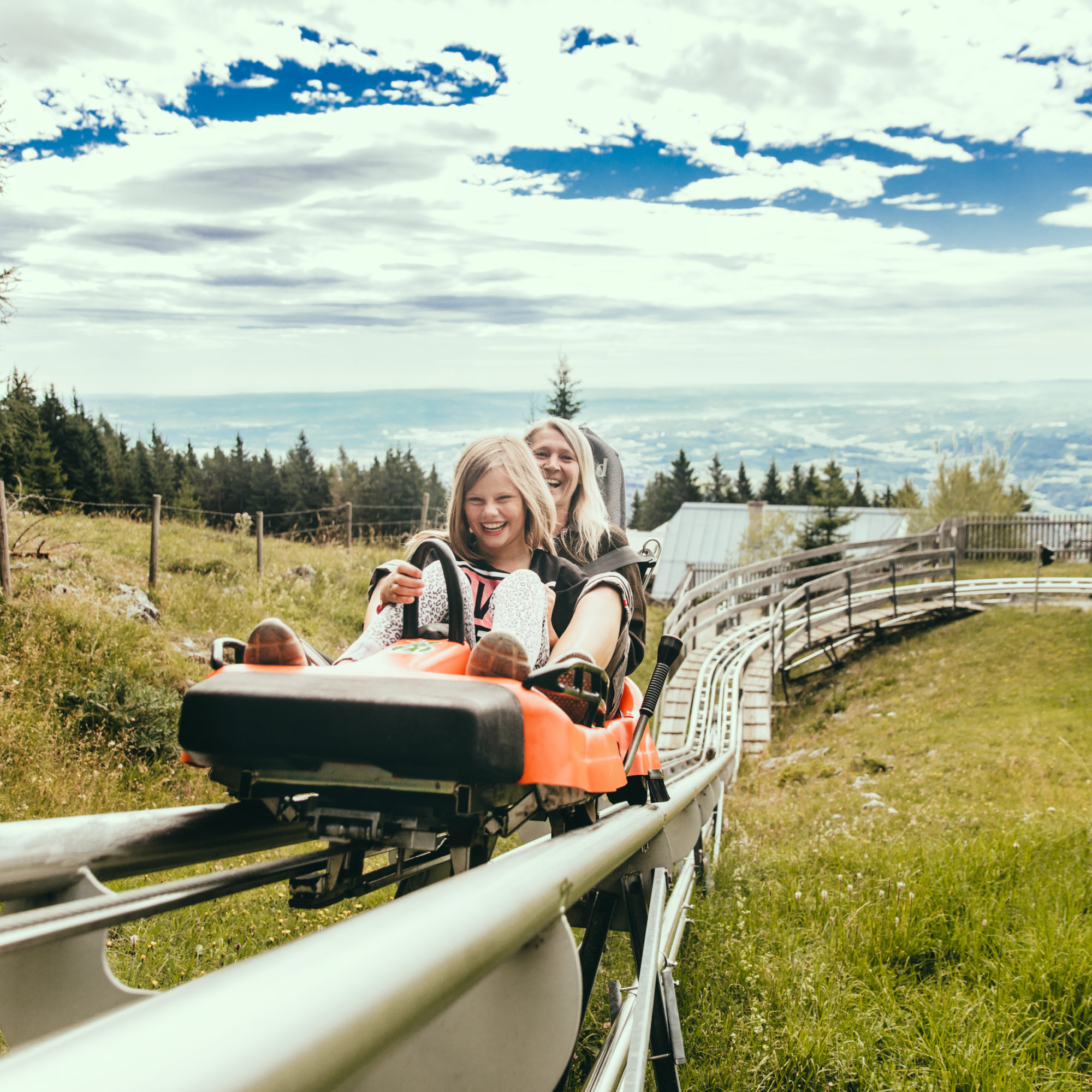 Two girls ride a mountain coaster uphill, smiling.