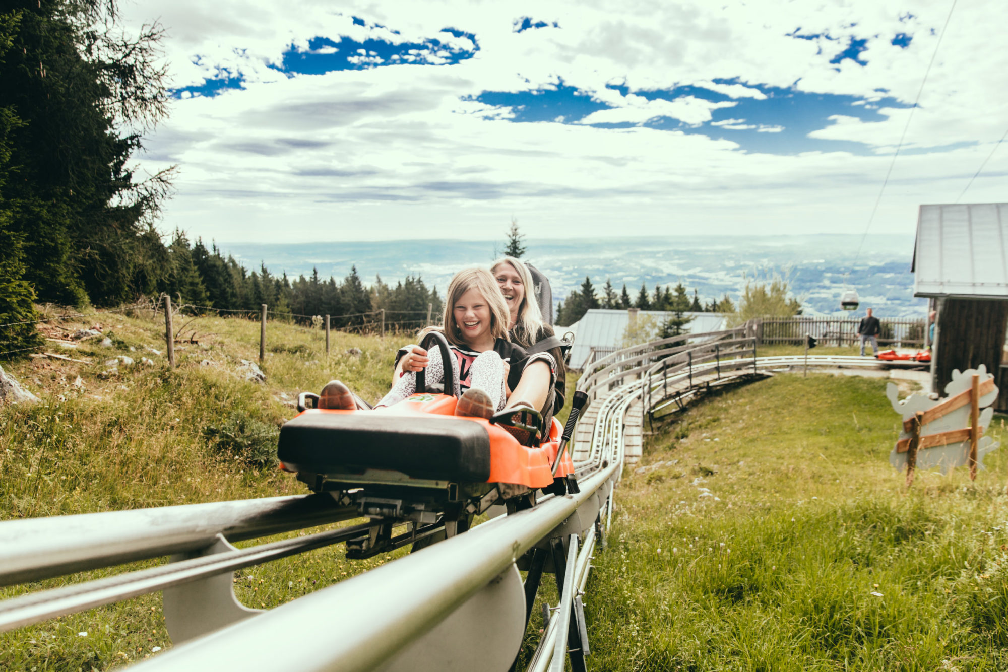Mädchen und Frau fahren mit dem Hexenexpress bergauf zur Bergstation. Sie lachen in die Kamera.