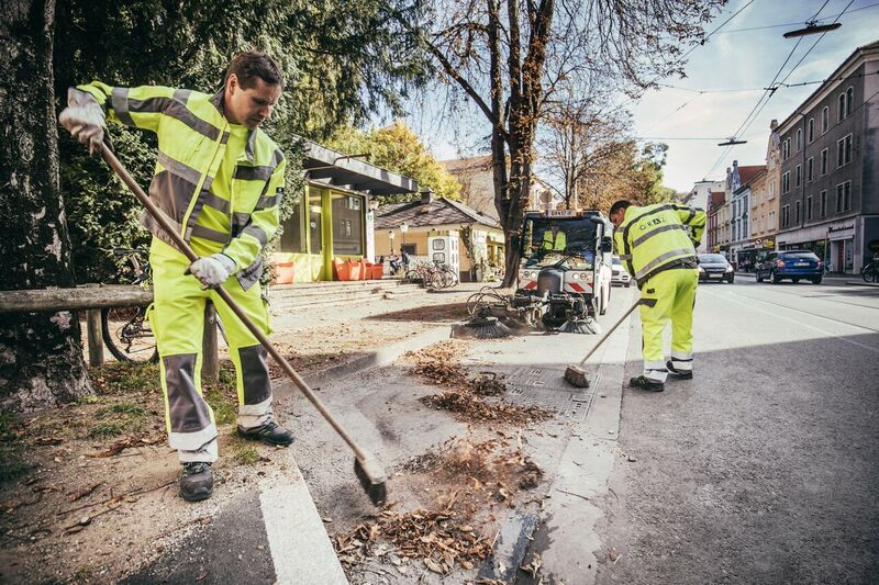 Zwei Holding Graz-Arbeiter in Arbeitskleidung kehren Laub und Schmutz am Straßenrand.