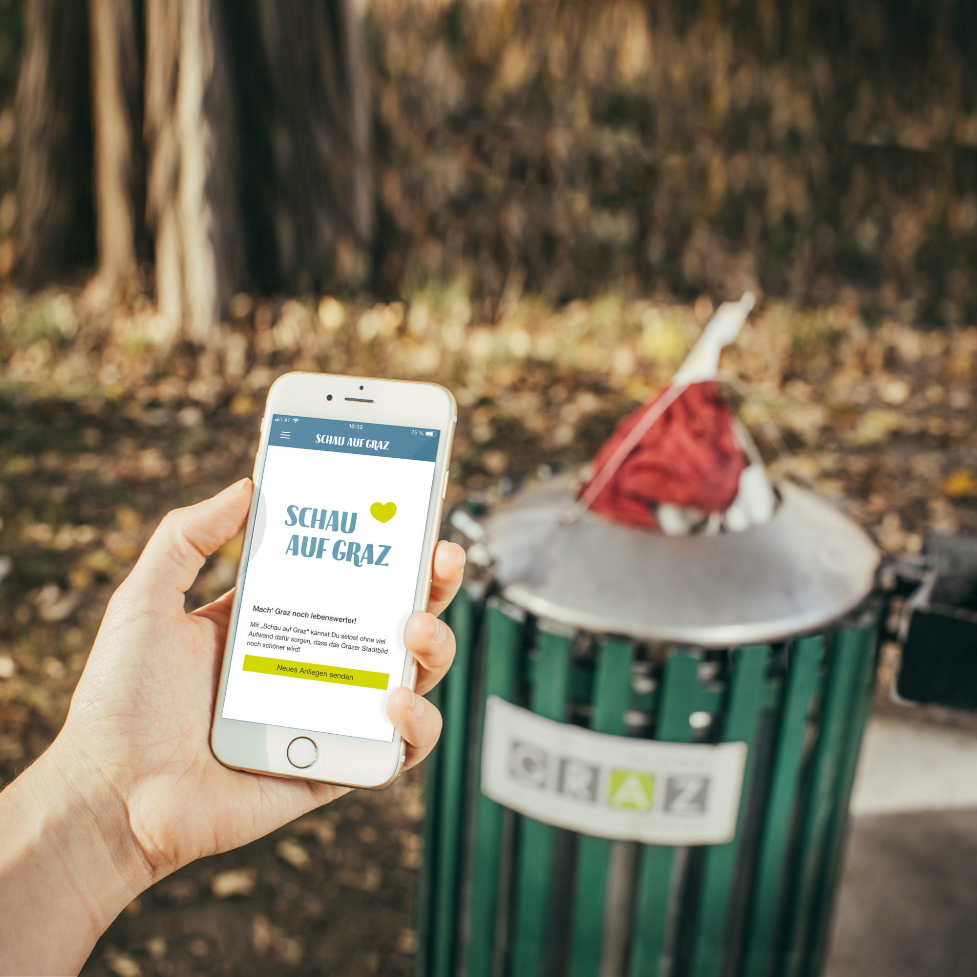Hand holding mobile phone with open 'Look at Graz' app of Holding Graz in front of rubbish bin in city park
