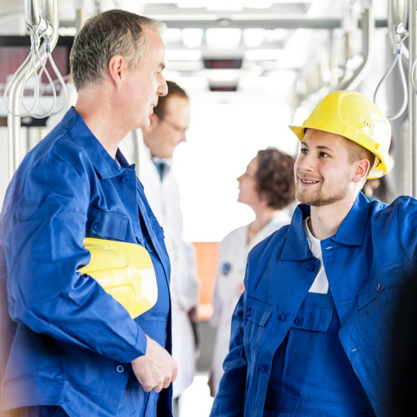 Zwei Arbeiter mit Helmen und in blauer Arbeitsmontur unterhalten sich während Straßenbahnfahrt.