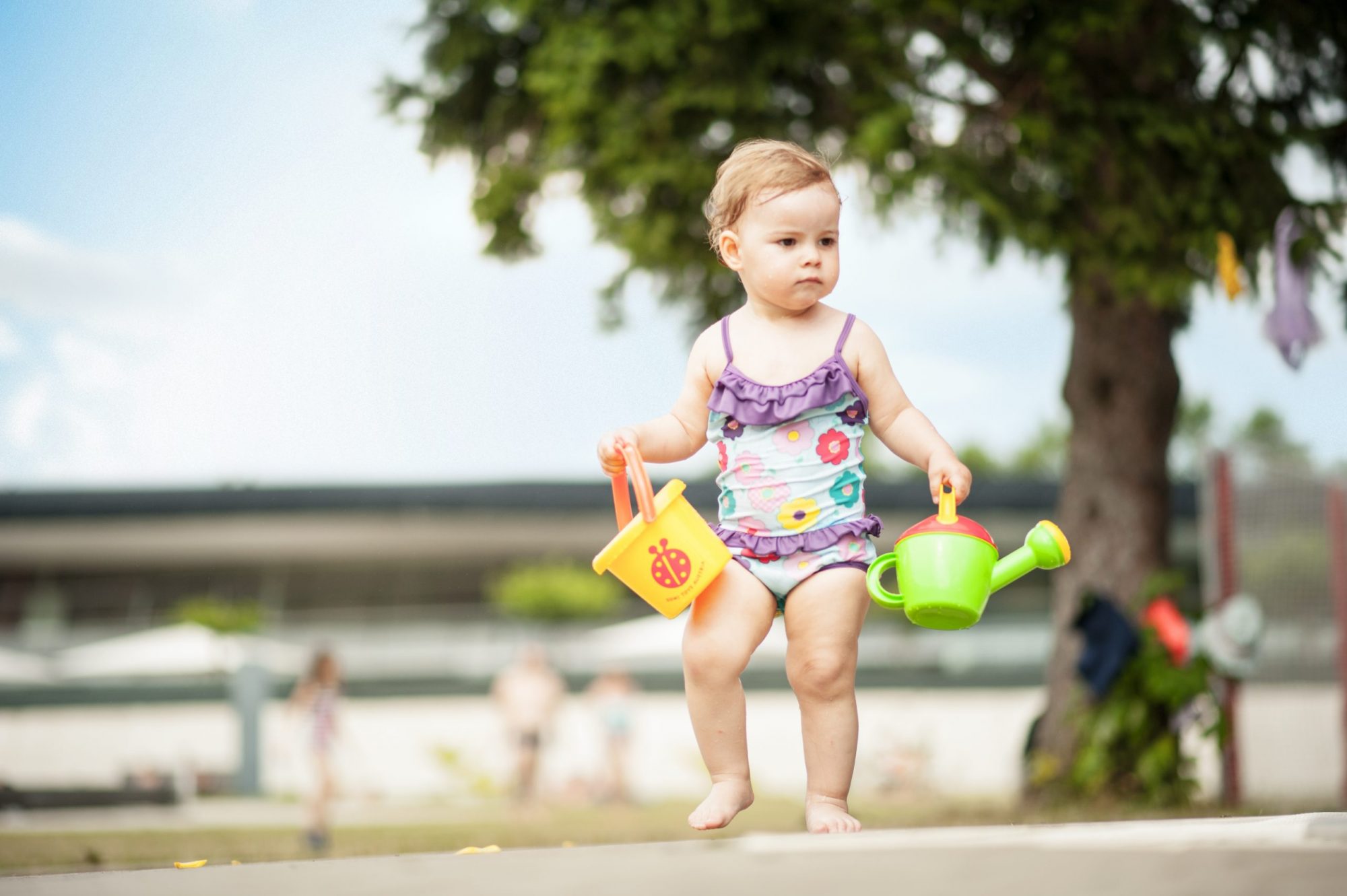A young girl in a swimsuit walks towards a children's pool, holding a yellow bucket and a green watering can.