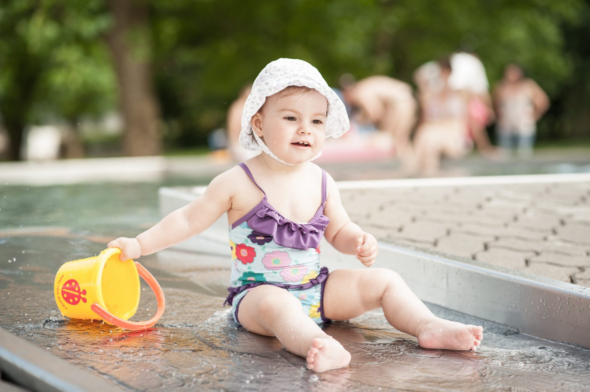 A young girl wearing a swimsuit and a sunhat plays with a yellow bucket in a shallow pool of water.