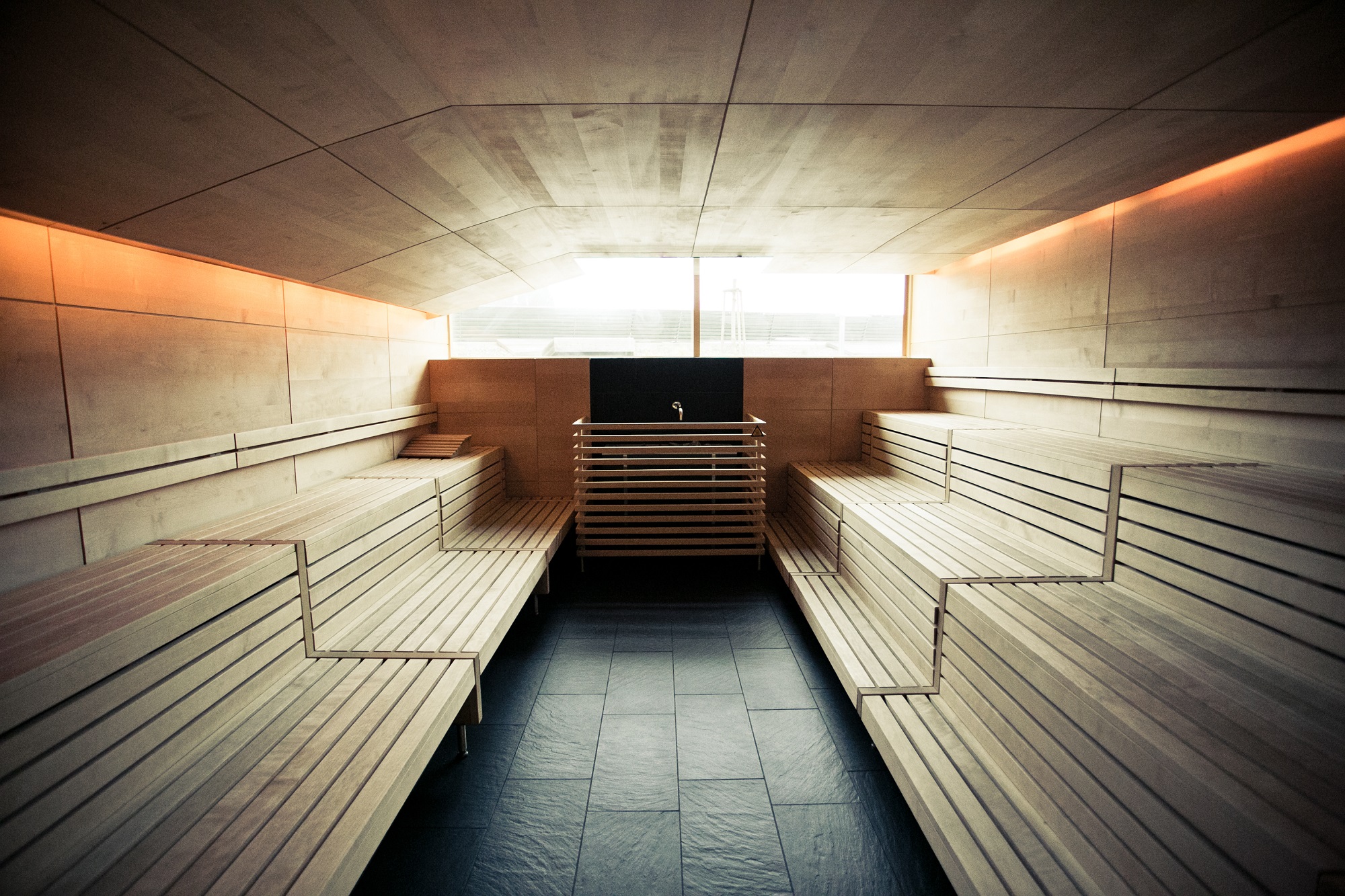 Empty sauna with wooden benches and a small sink in the middle. The room is lit with a warm, orange glow.