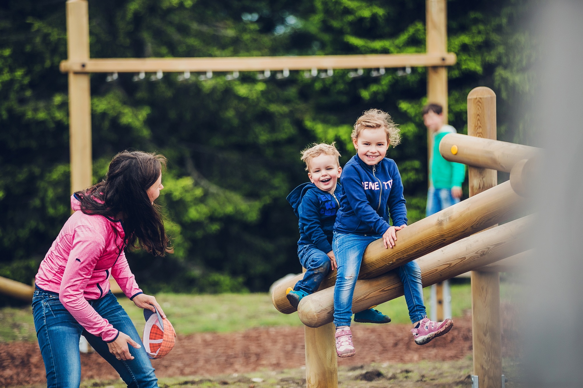 A woman watches two children play on a wooden balance beam at the Schöckl motor skills parkour. "GENE TY" is printed on the girl's sweatshirt.