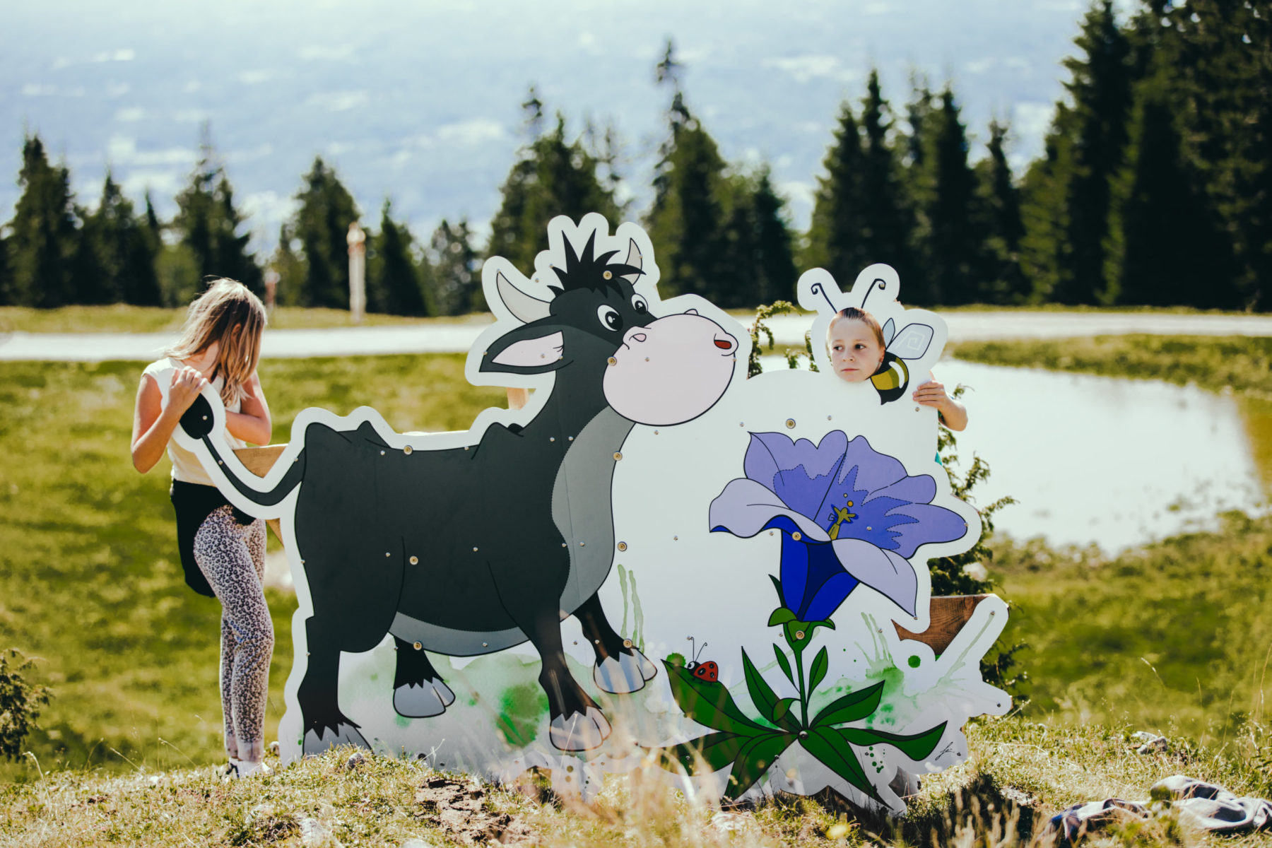 Zwei Kinder posieren mit Kuh-Skulptur. Mädchen hält Schwanz der Kuh, Bub streckt Kopf durch Loch.
