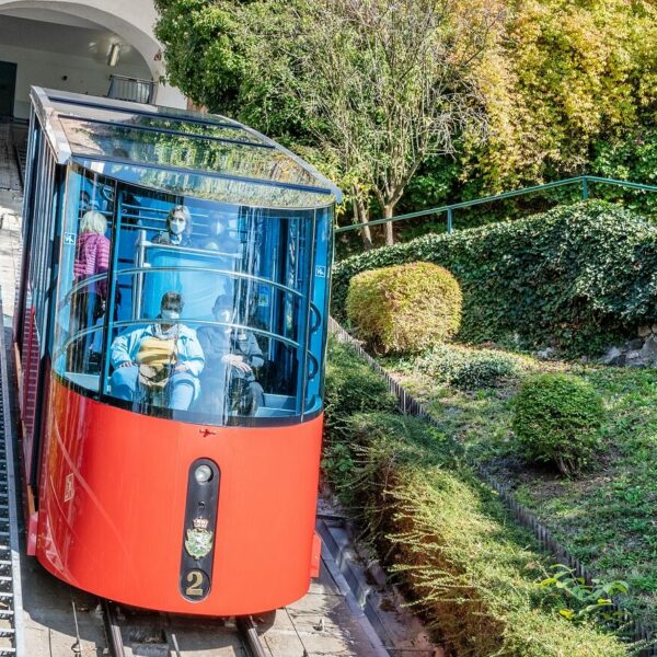 Moderne rote Garnitur der Schloßbergbahn mit Glasdach fährt mit Passagieren durch Hangbereich.