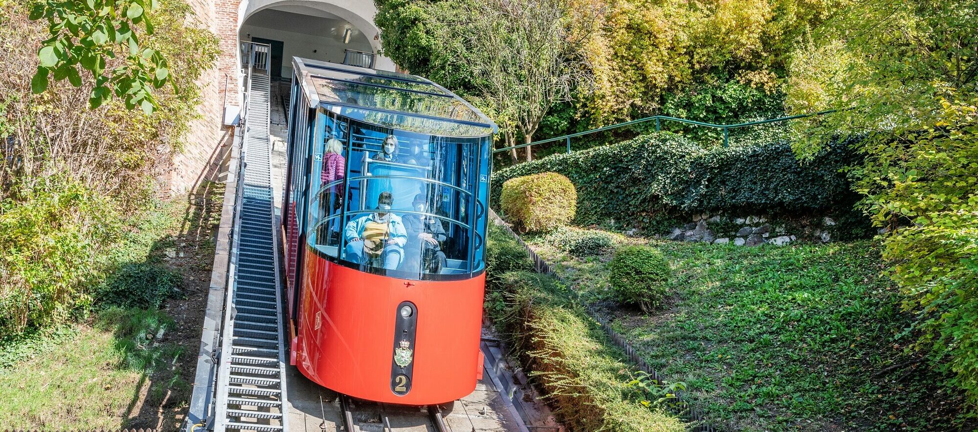 Moderne rote Garnitur der Schloßbergbahn mit Glasdach fährt mit Passagieren durch Hangbereich.