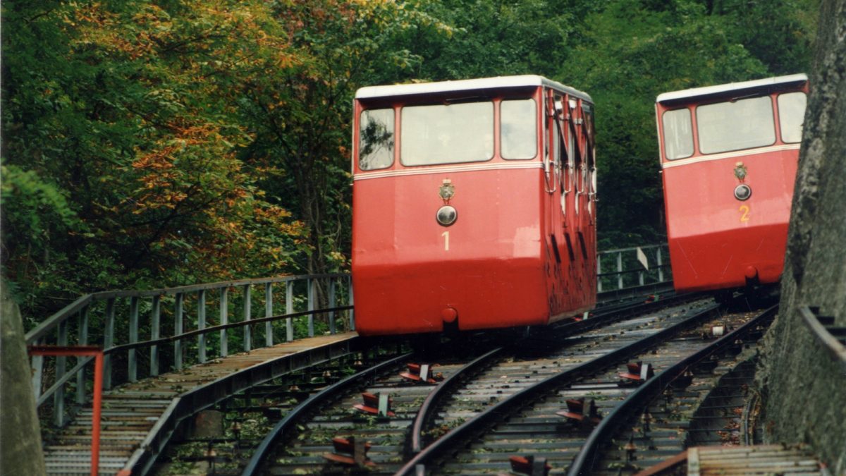 Two red funicular cars, numbered 1 and 2, ascend a track through a wooded area. The Schlossbergbahn in Graz, Austria.
