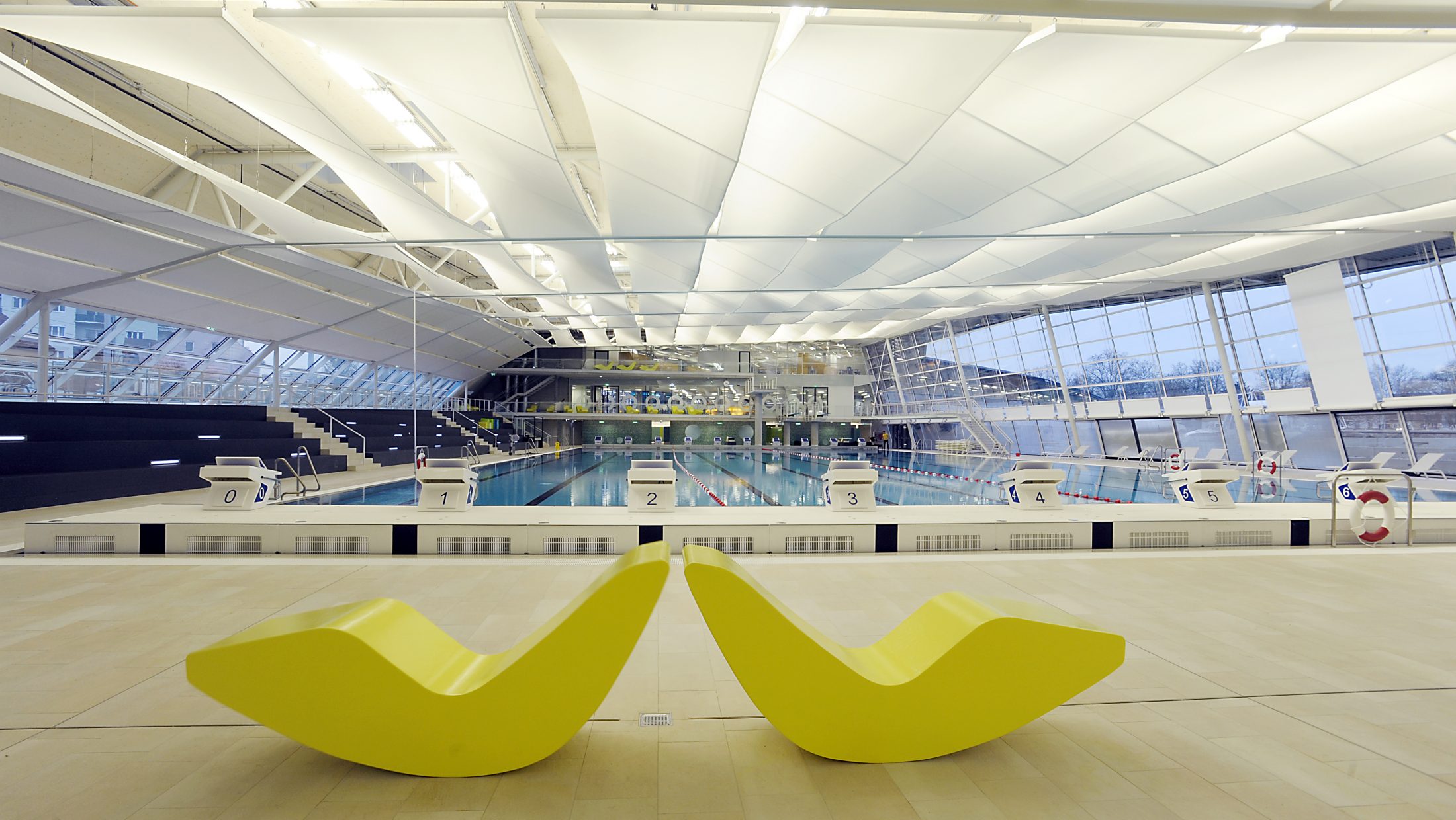 Auster Sportbad: Innenansicht mit Schwimmbahnen, Sprungbrettern und gelben Liegestühlen im Vordergrund.