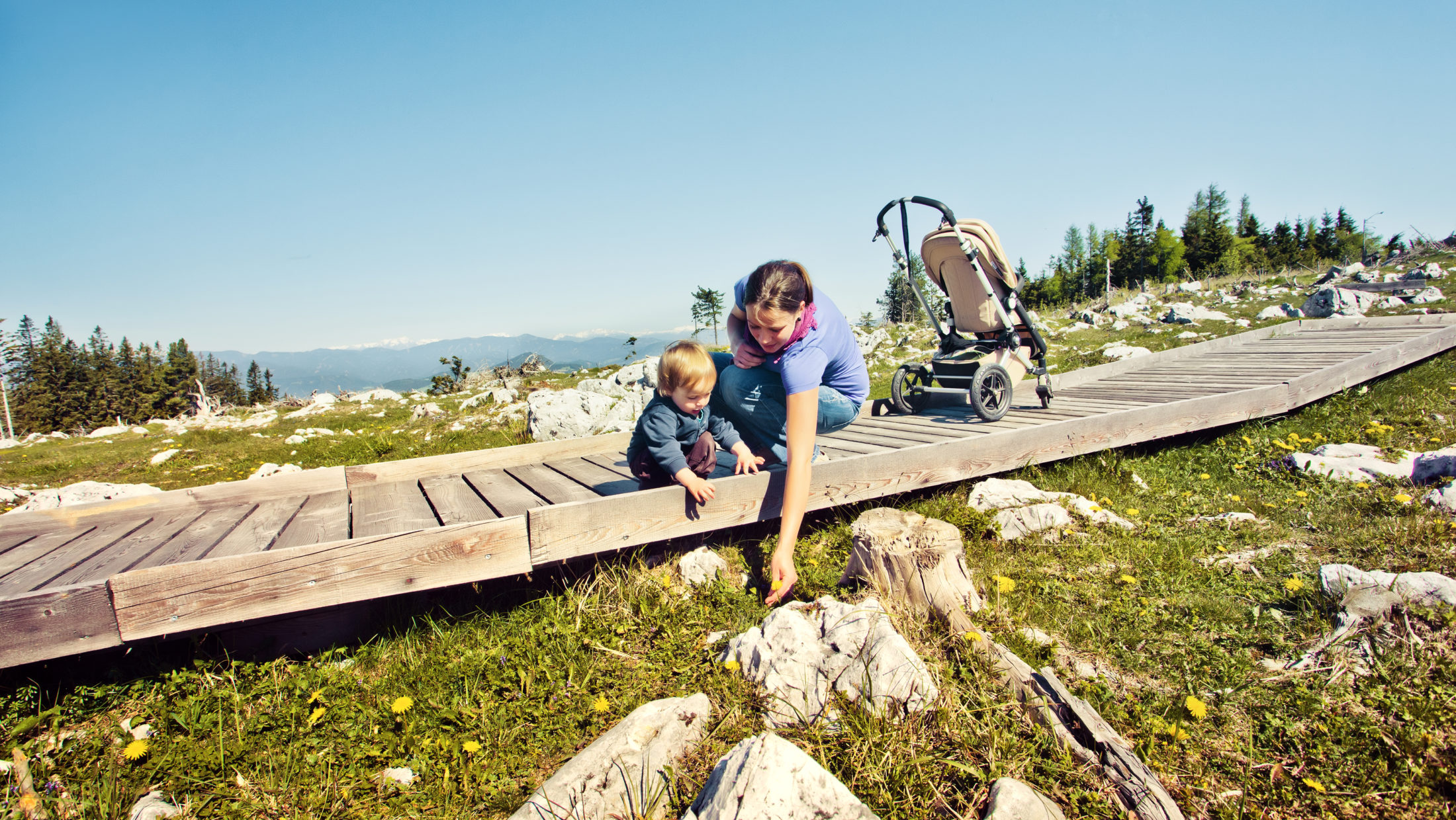 A mother and her toddler are on a wooden pathway on the Schöckl plateau, with a stroller nearby.