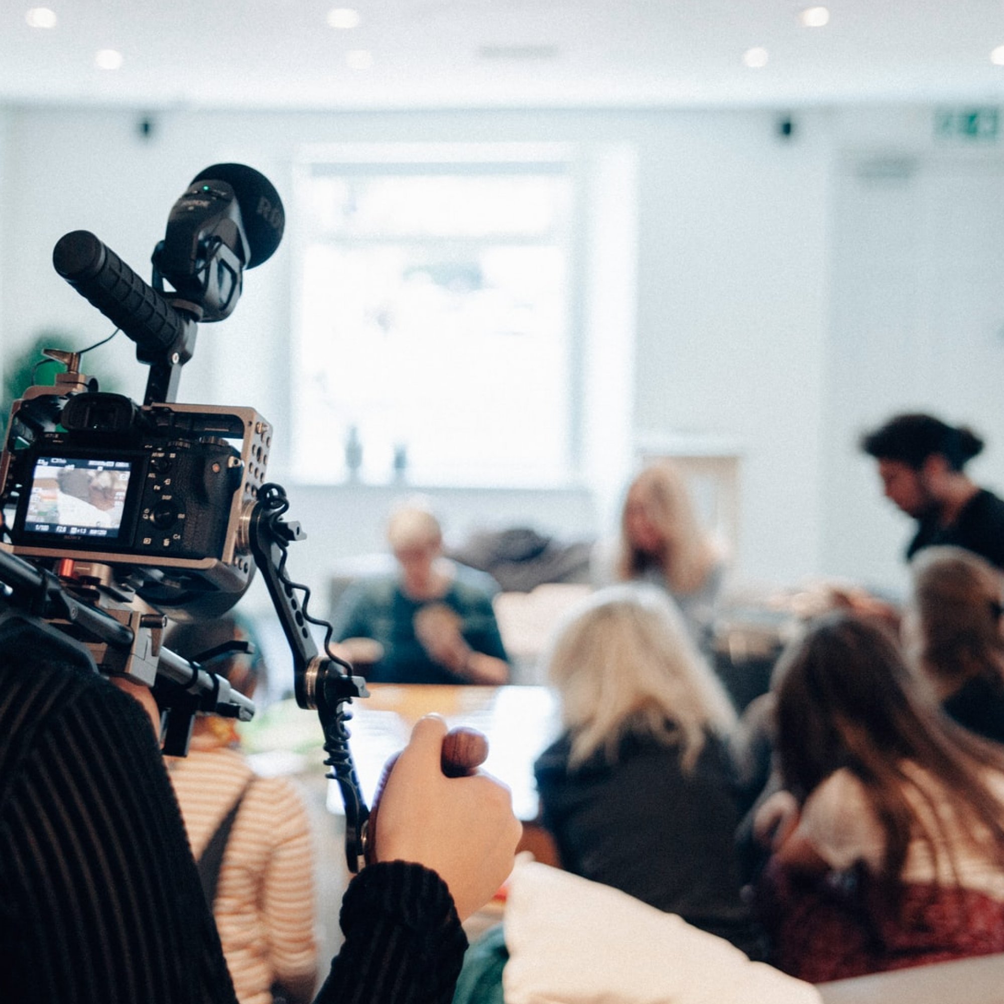 Frau mit Kamera filmt eine Gruppe von Menschen in einem hellen Raum, Fokus liegt auf Kamera.