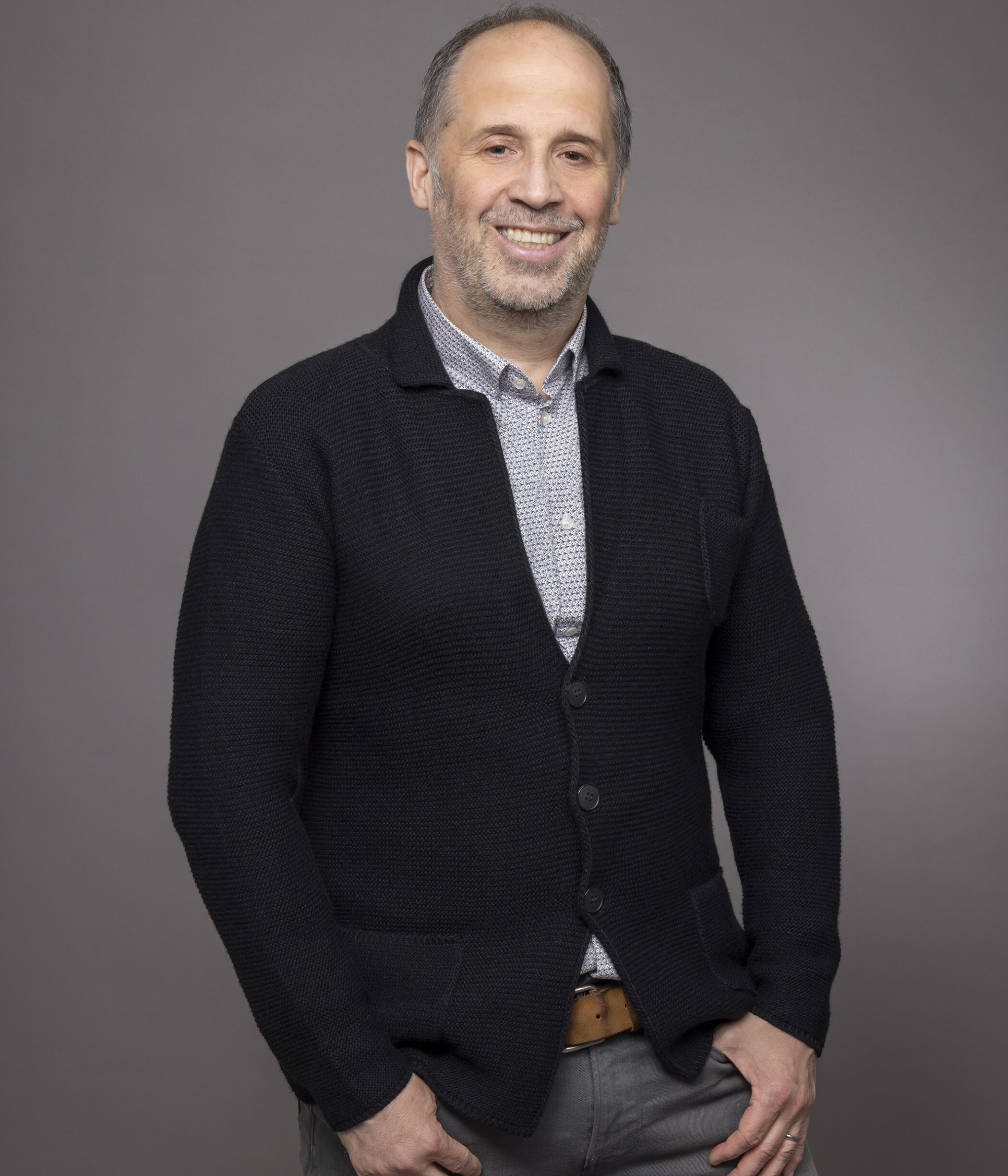 Man smiles wearing button-down shirt, cardigan, jeans, & rings.