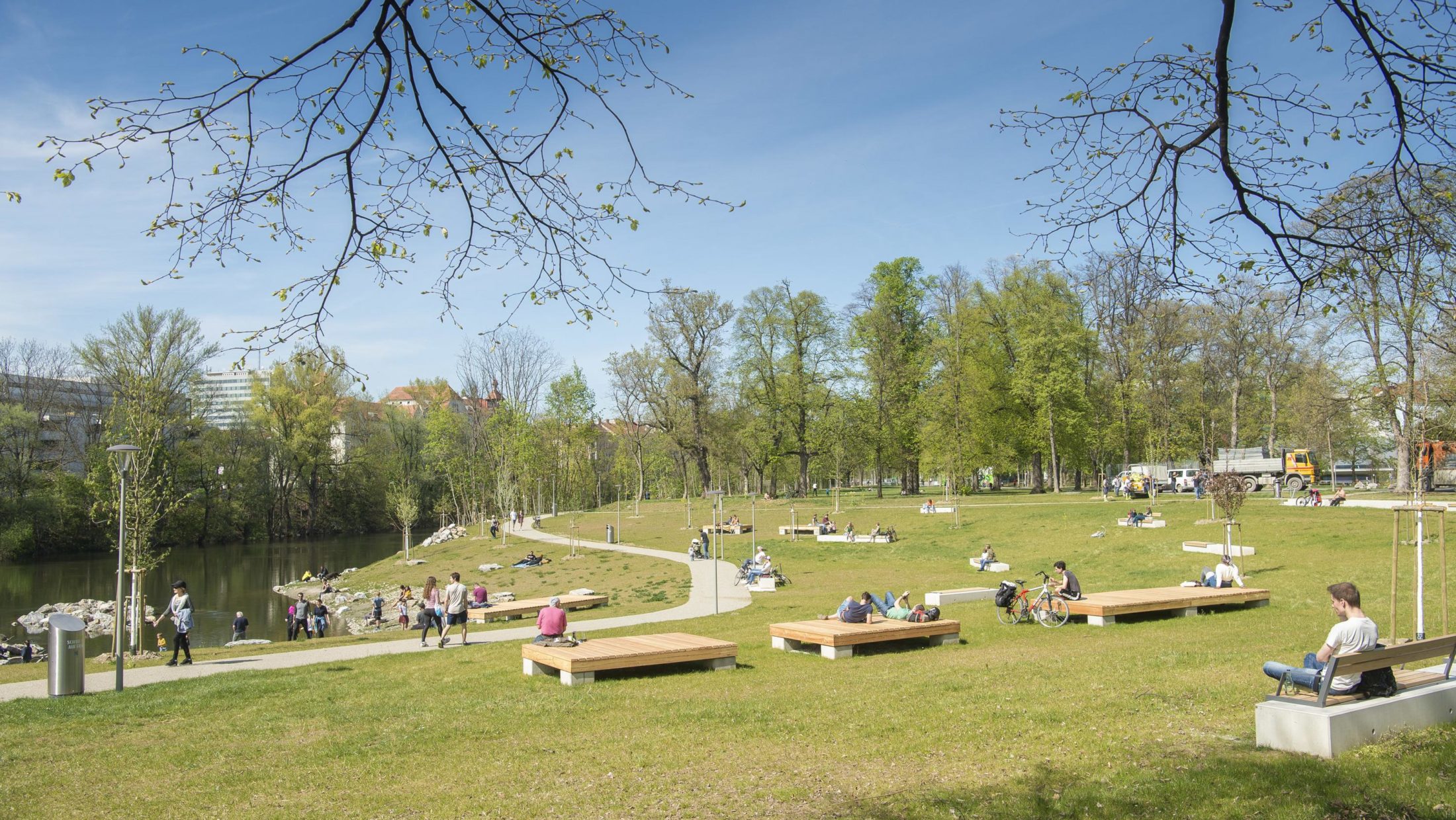 A sunny park scene with people relaxing on wooden benches. A small pond in the foreground.