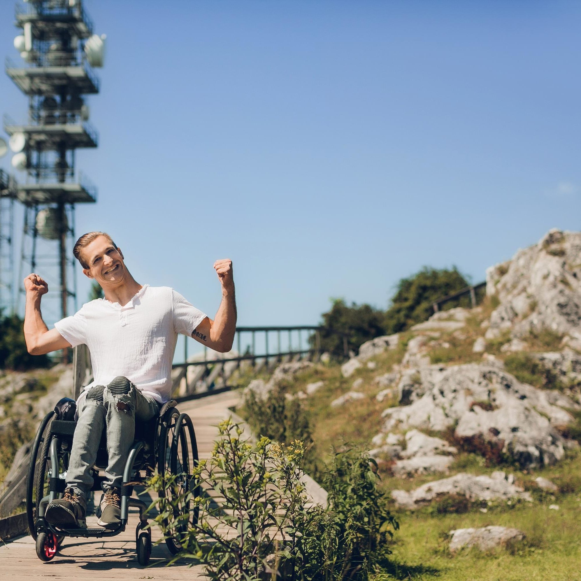 A man in a wheelchair smiles and raises his arms in celebration on a paved path near the Schöckl.