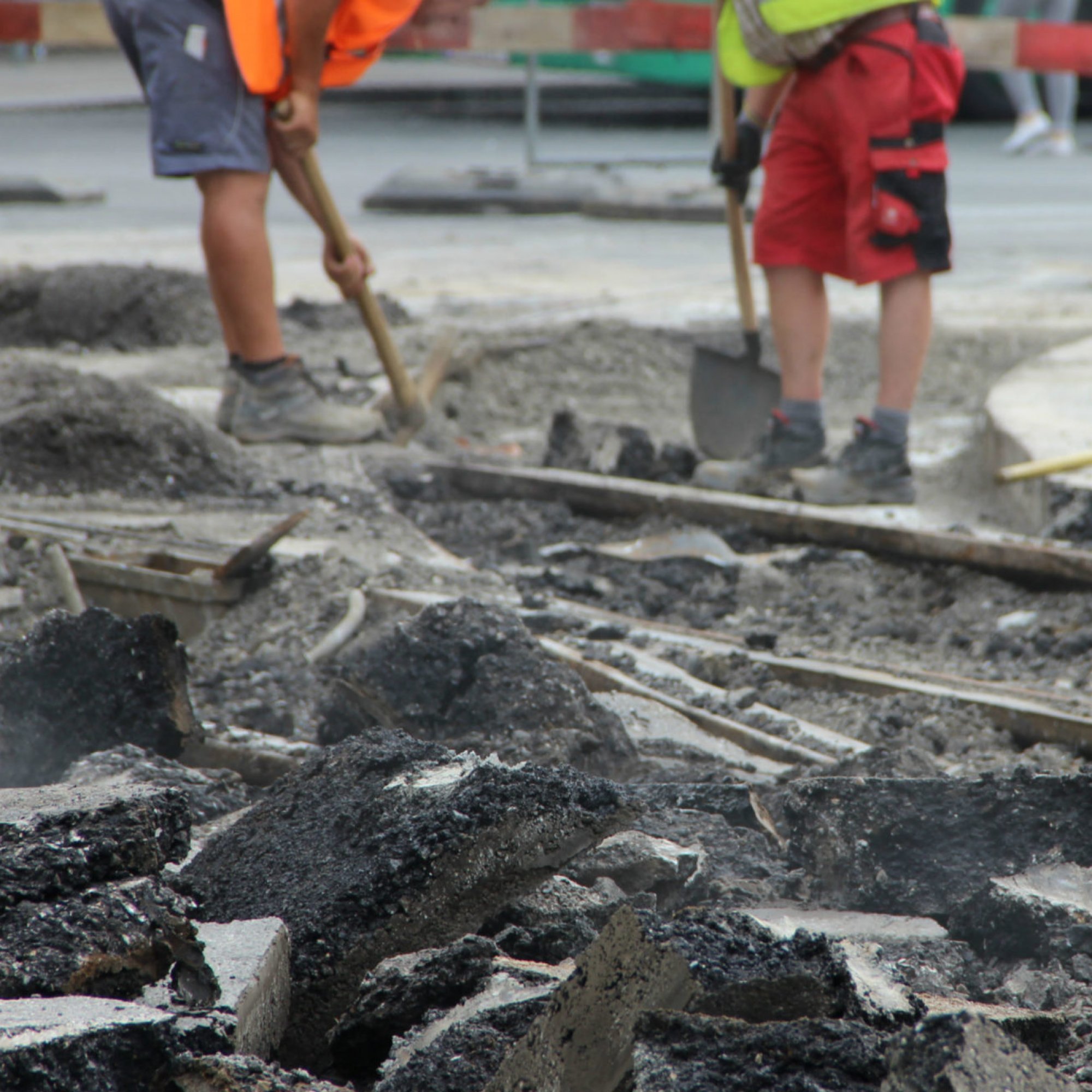 Zwei Bauarbeiter mit Schaufel und Spitzhacke bei Gleisbauarbeiten, vorne aufgebrochener Asphalt.