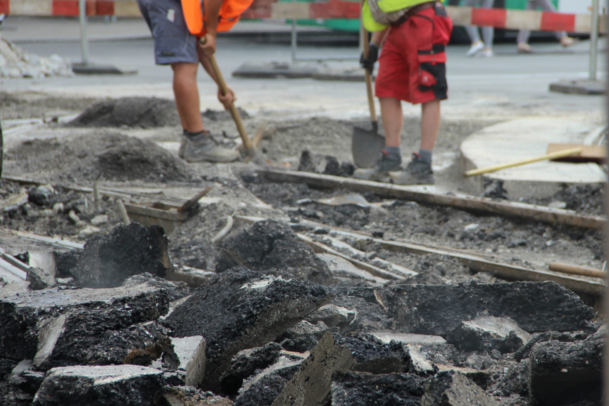 Zwei Bauarbeiter mit Schaufel und Spitzhacke bei Gleisbauarbeiten, vorne aufgebrochener Asphalt.