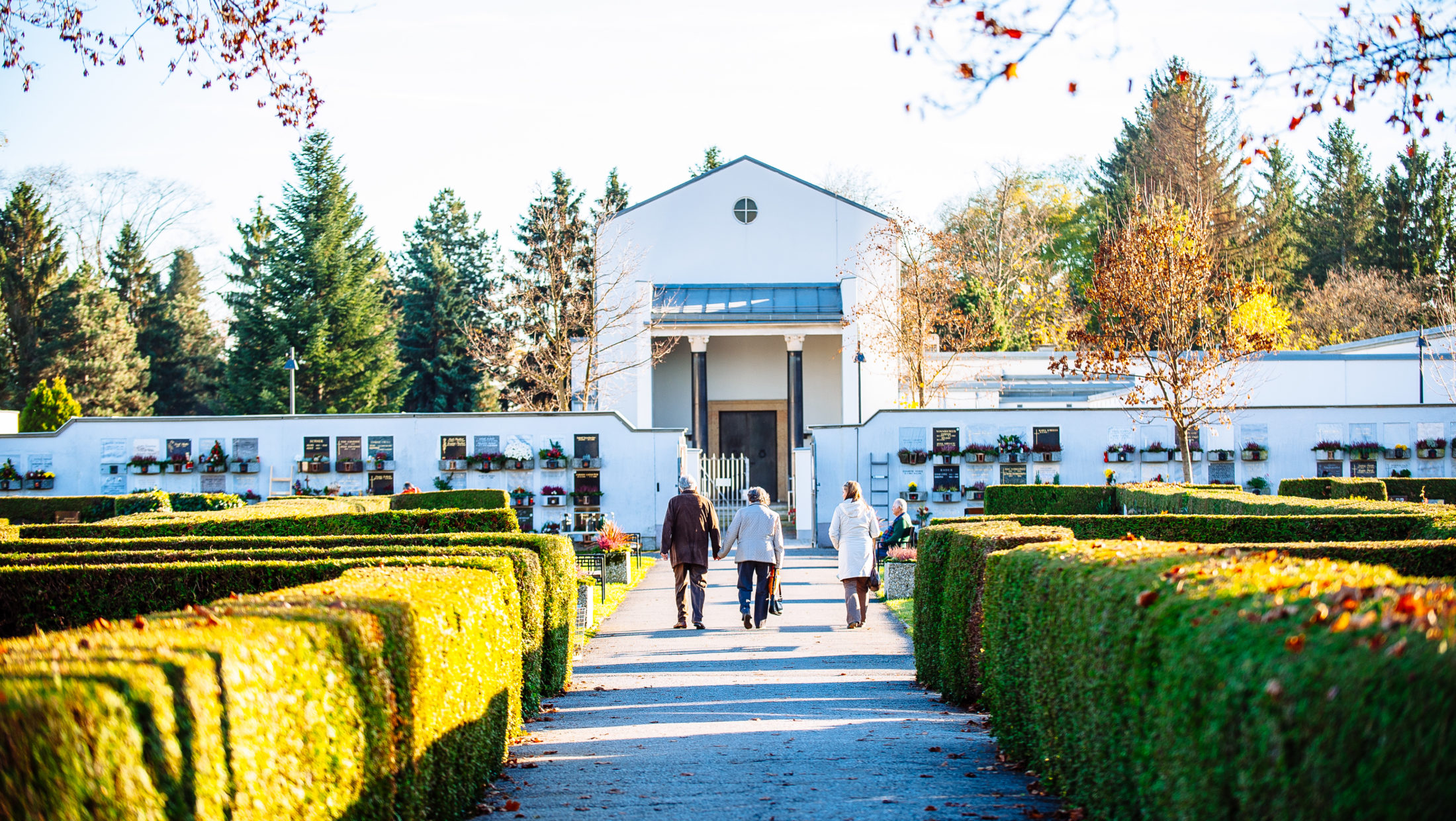 Three people walk along a path towards the ceremonial hall of the Graz Urn Cemetery.