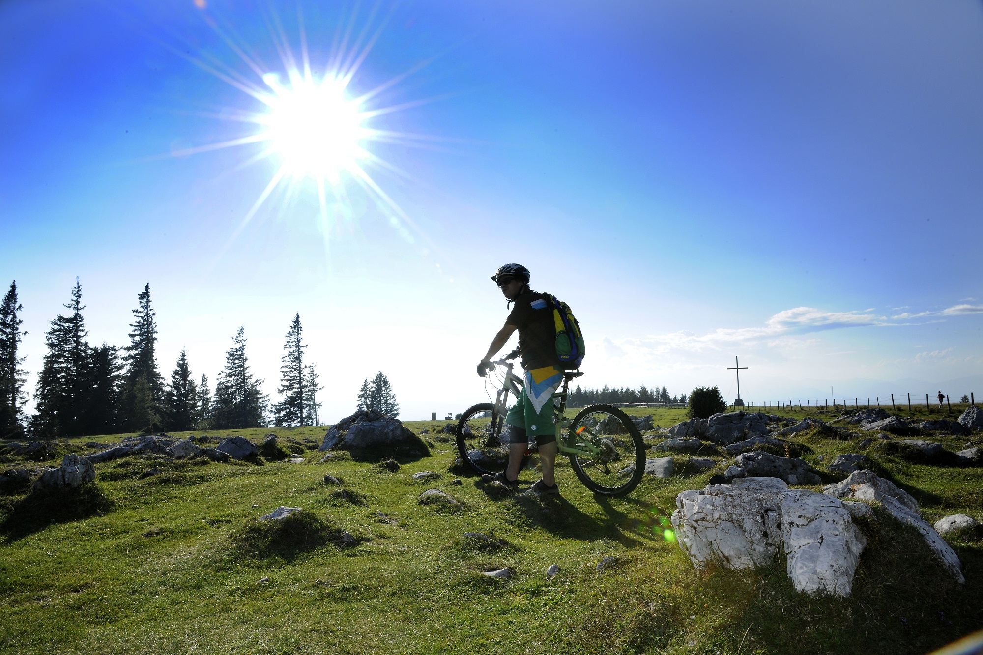 A mountain biker stands on a grassy hillside with a bright sun shining above. The Schöckl mountain is in the background.