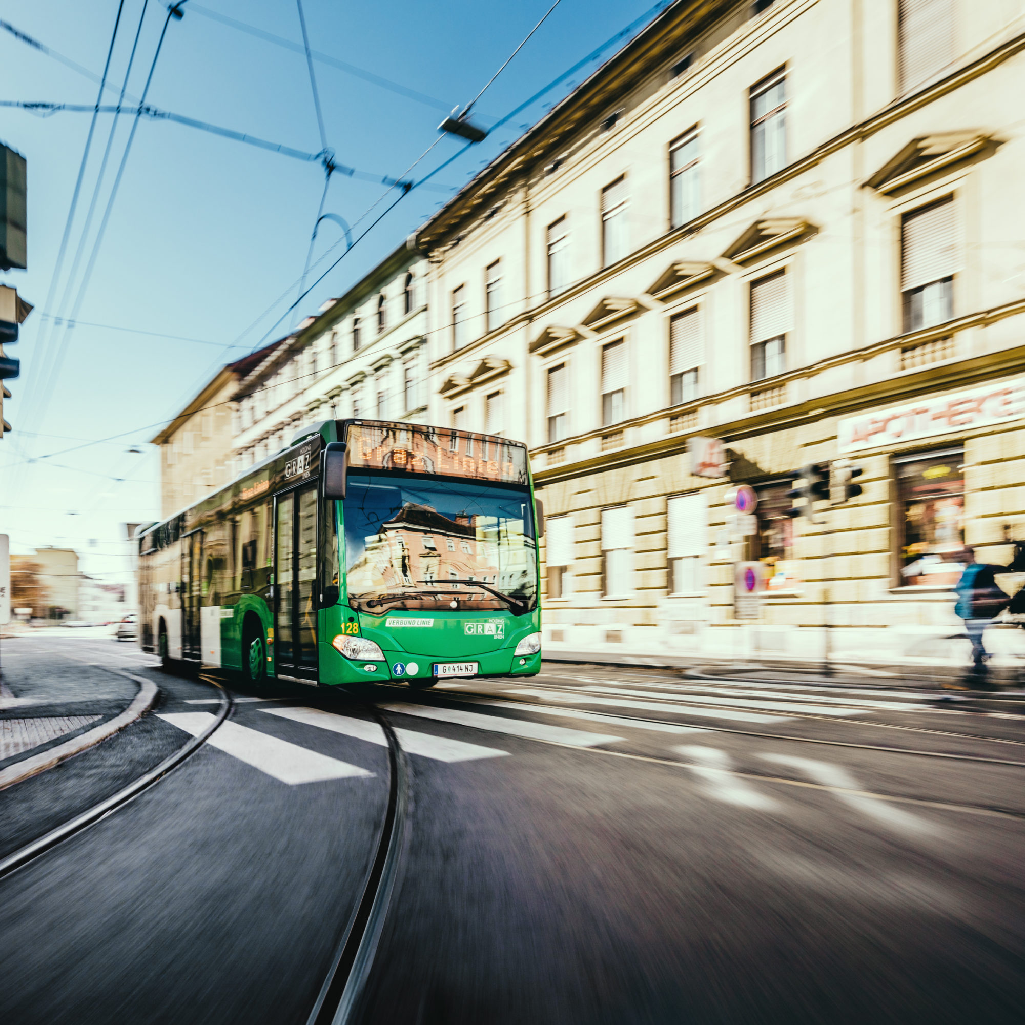 A green Graz Linien bus turns a corner at an intersection in the city. The bus is blurred due to motion.