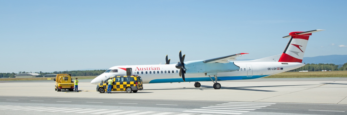An Austrian Airlines plane is parked at Graz Airport with airport staff nearby. The plane's registration number is OE-LGH.