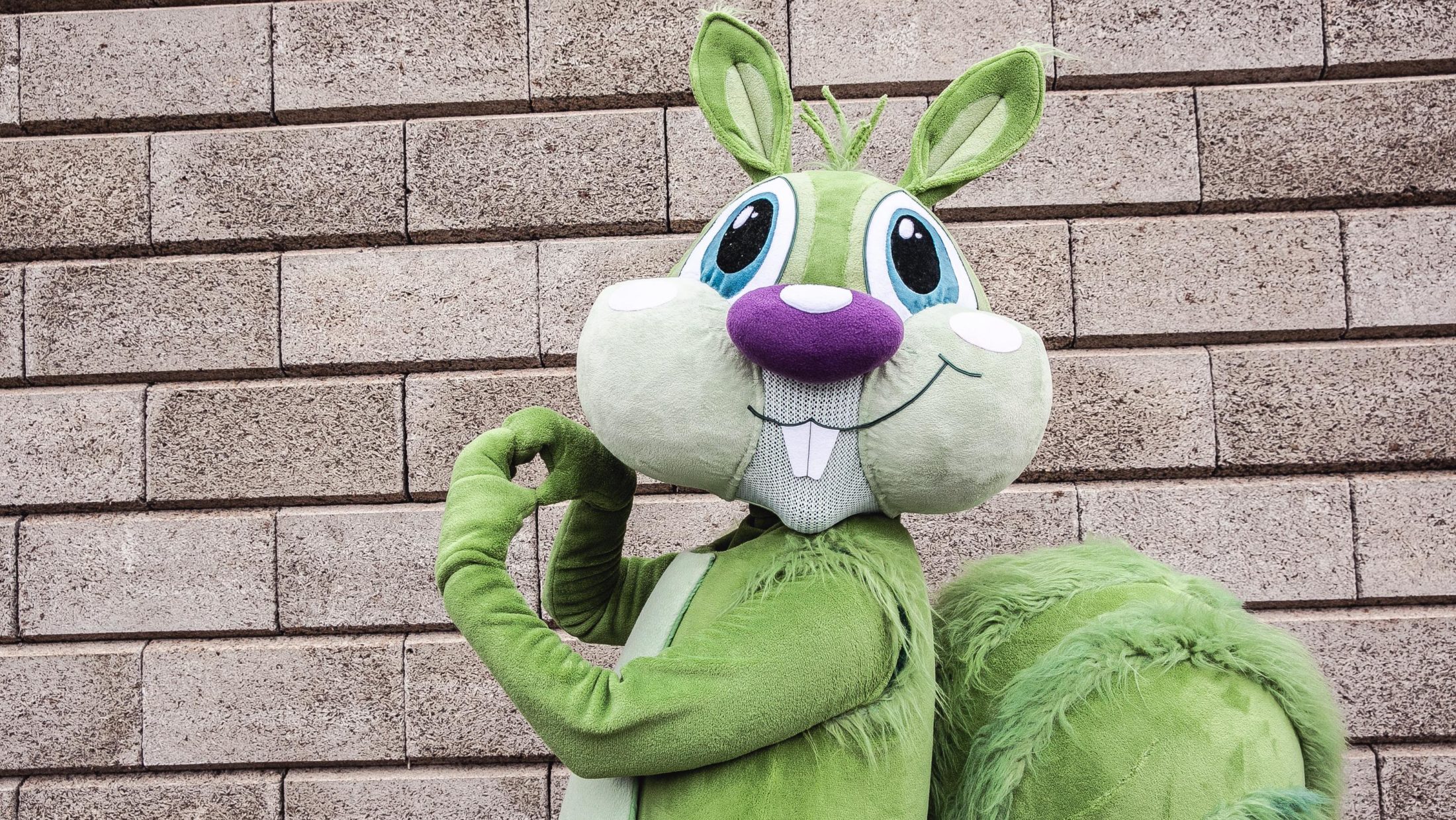 The Graz mascot Hansi, a green squirrel, stands in front of a brick wall and makes a heart shape with his paws.