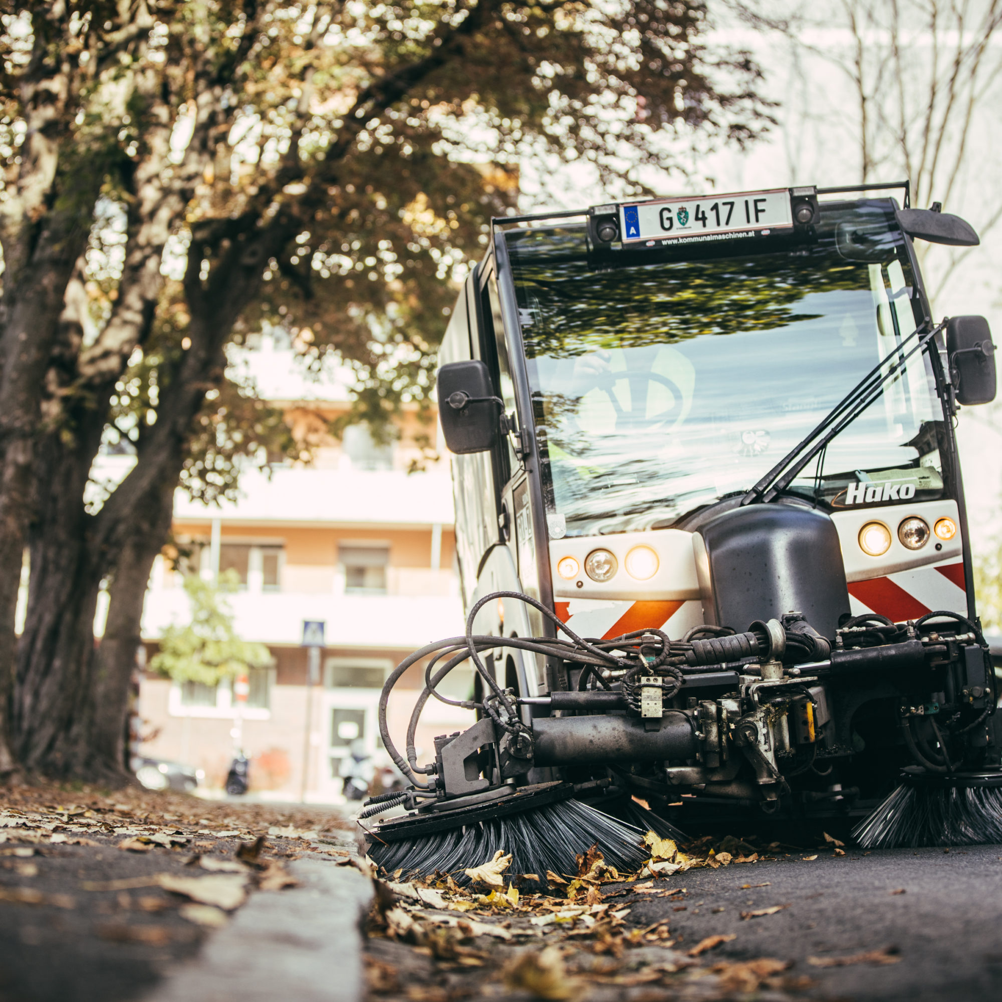 Kehrmaschine reinigt eine von Herbstlaub bedeckte Straße in einer urbanen Umgebung.
