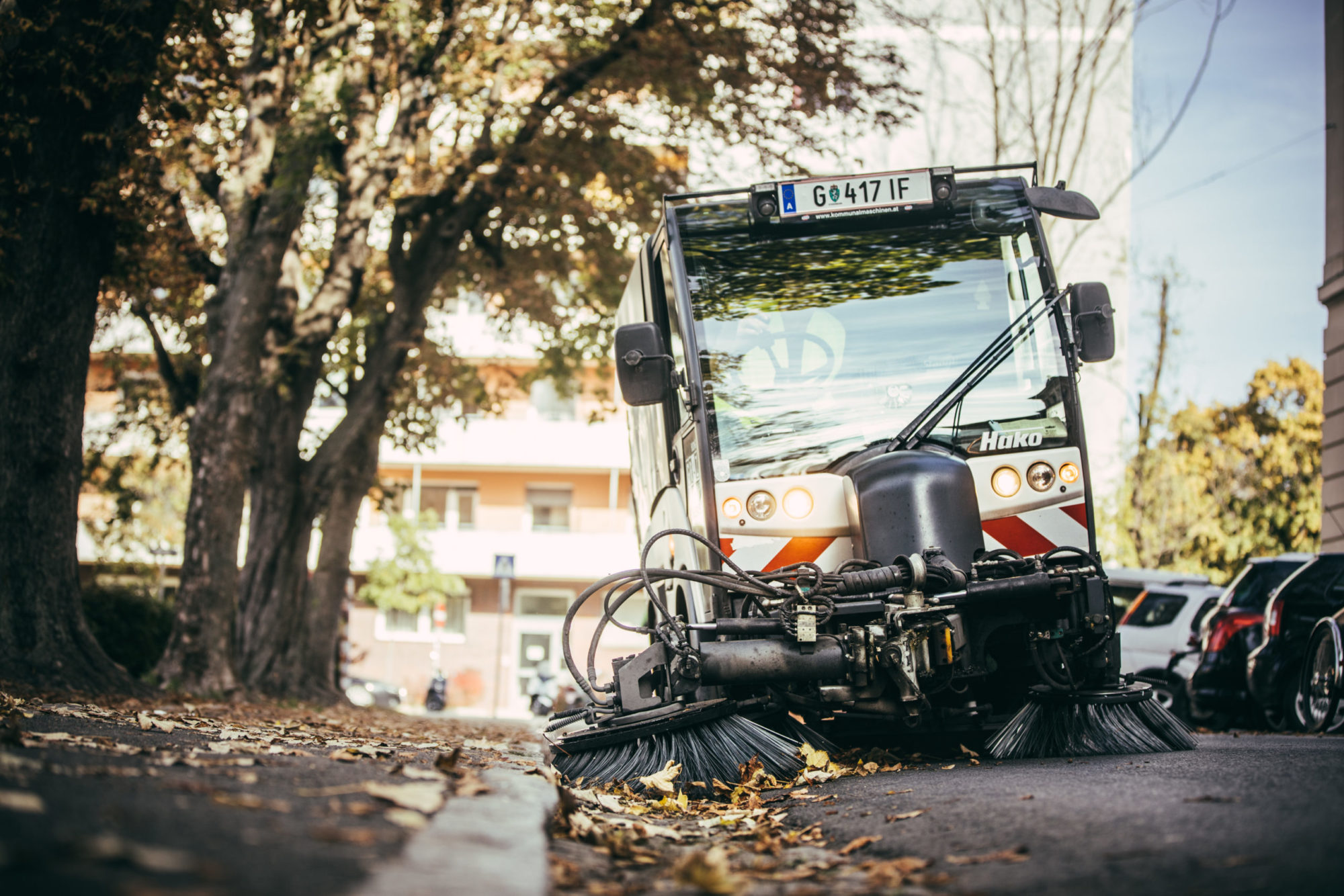Kehrmaschine reinigt eine von Herbstlaub bedeckte Straße in einer urbanen Umgebung.