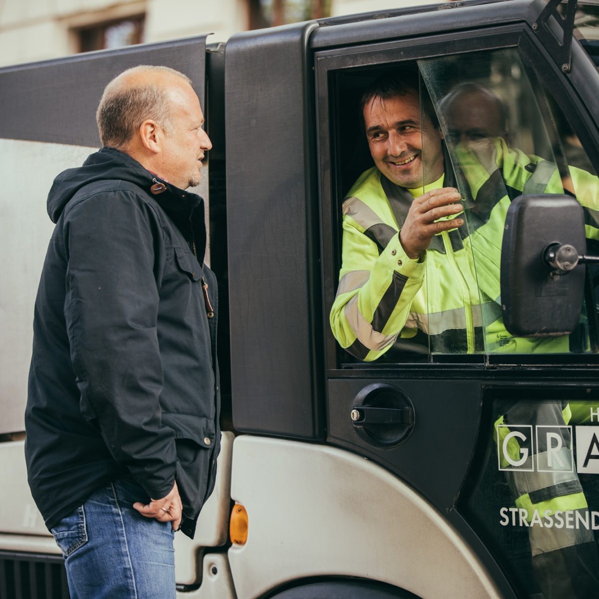 A man talks to a worker in a yellow vest sitting in a small white truck marked "Holding Graz Strassendienst."