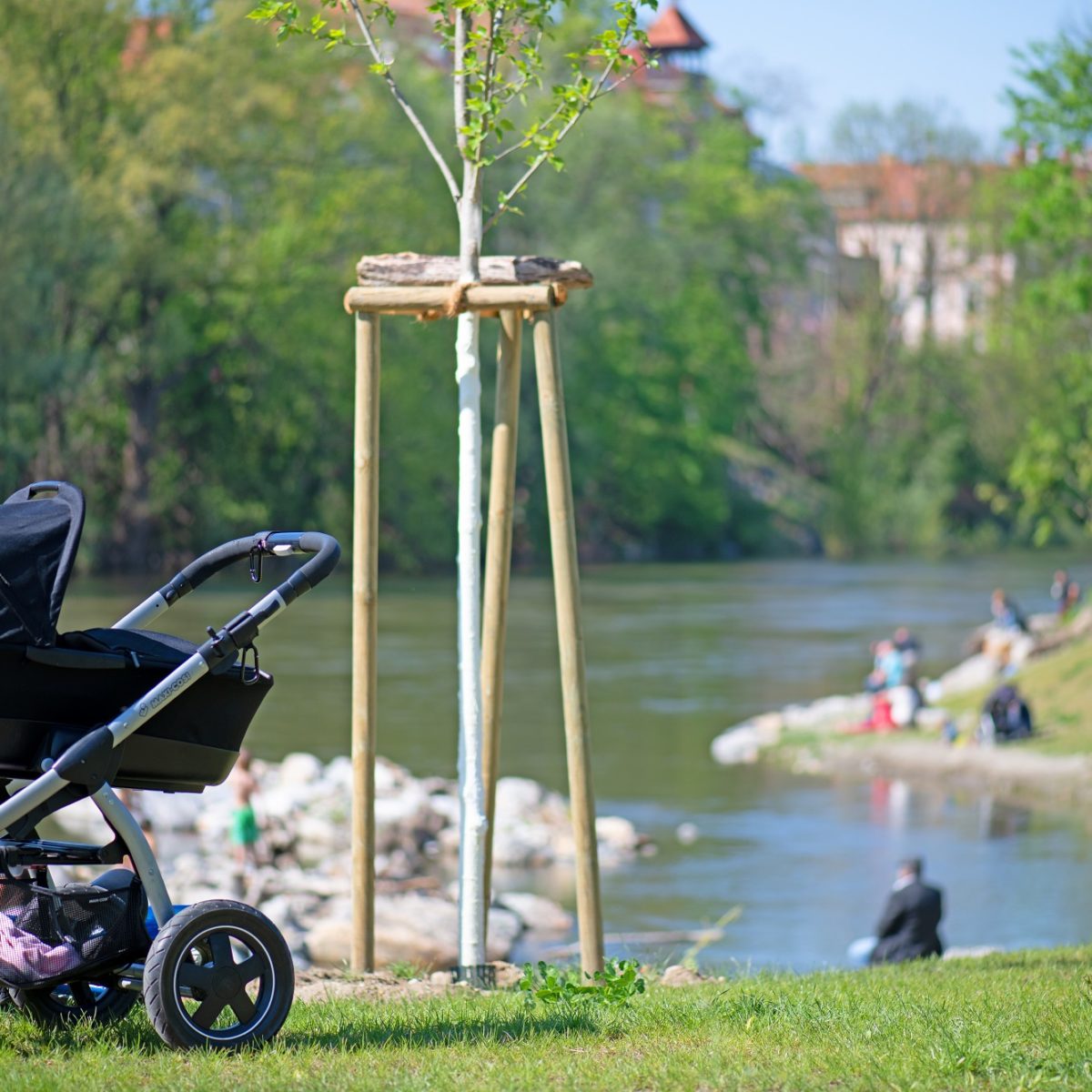 A stroller sits on a grassy bank overlooking a river with people sitting on the opposite bank.