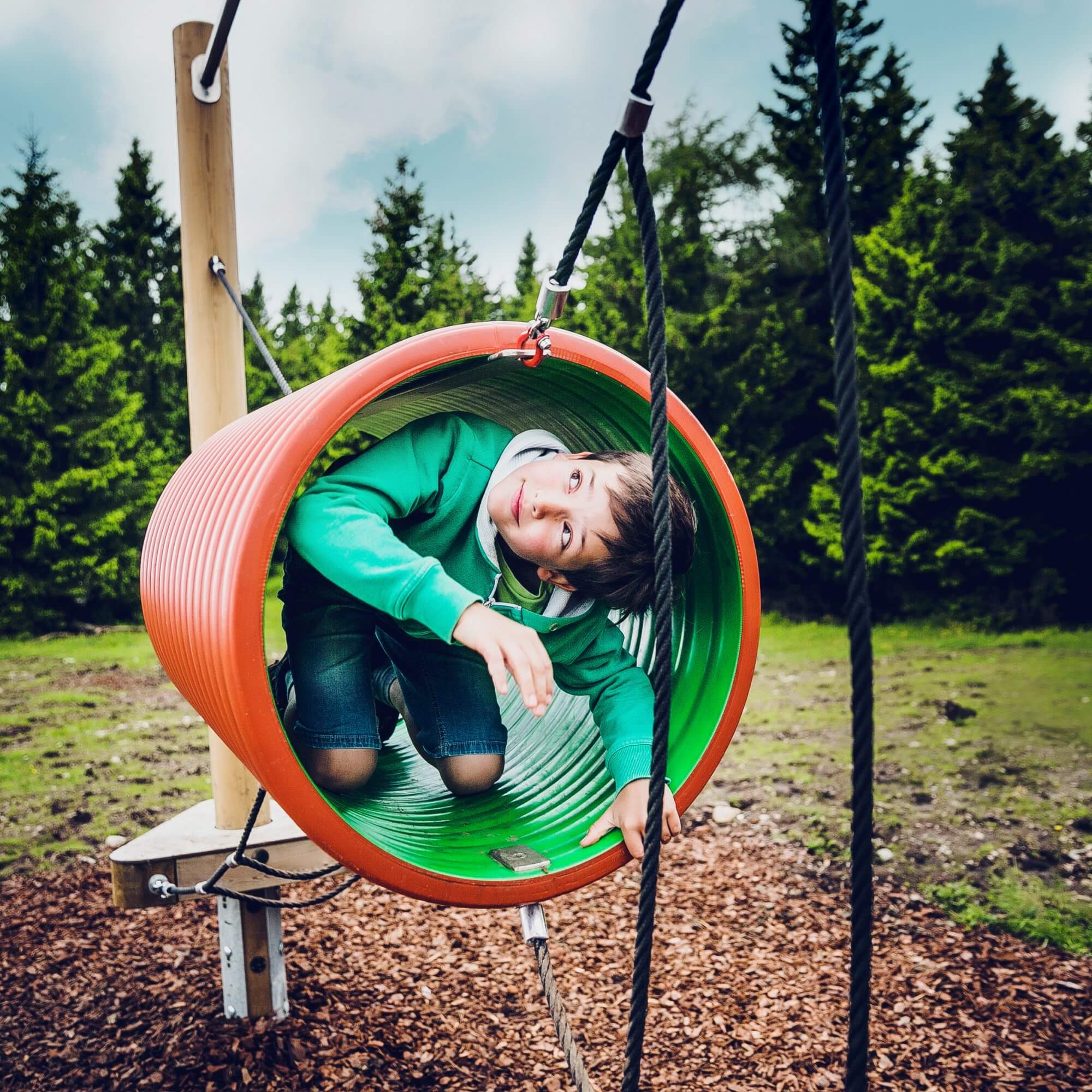 A young boy crawls through a green and orange tube at a playground on the Schöckl mountain.