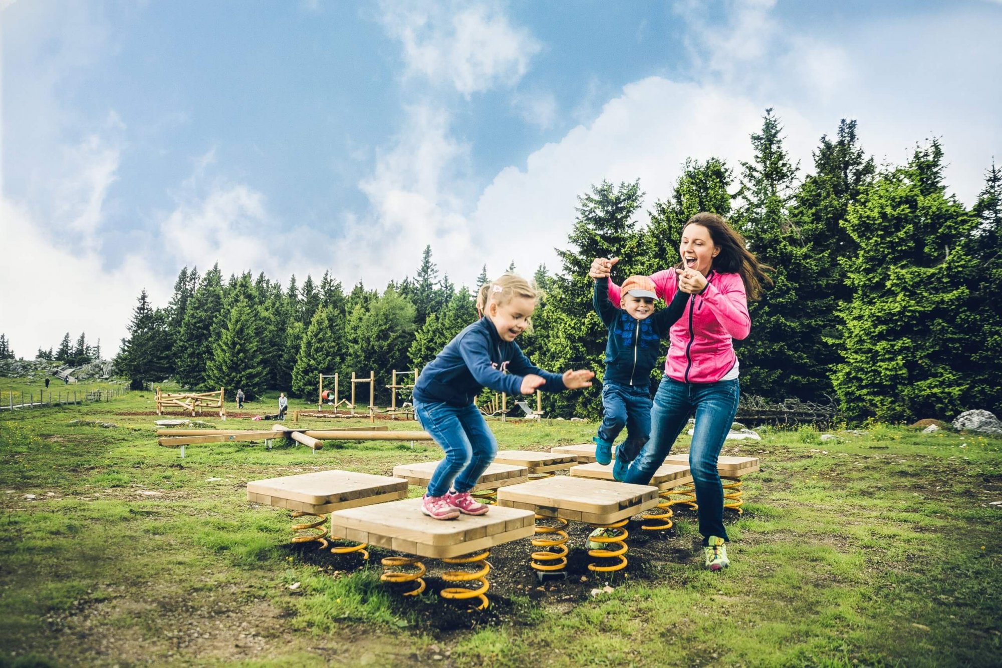 Mutter mit Bub und Mädchen hüpfen auf gefederten Holzplatten im Motorikparkour Schöckl