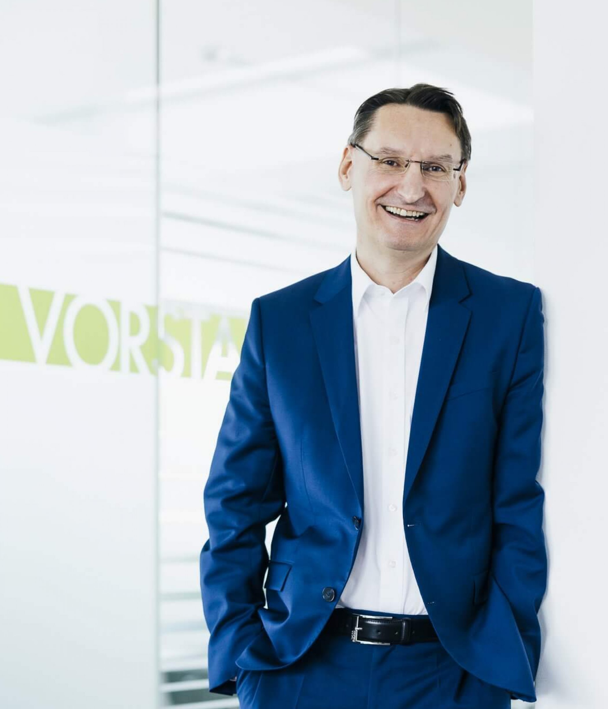 Gert Heigl, wearing a blue suit, smiles while leaning against a wall. Behind him, a sign reads "Vorstand".