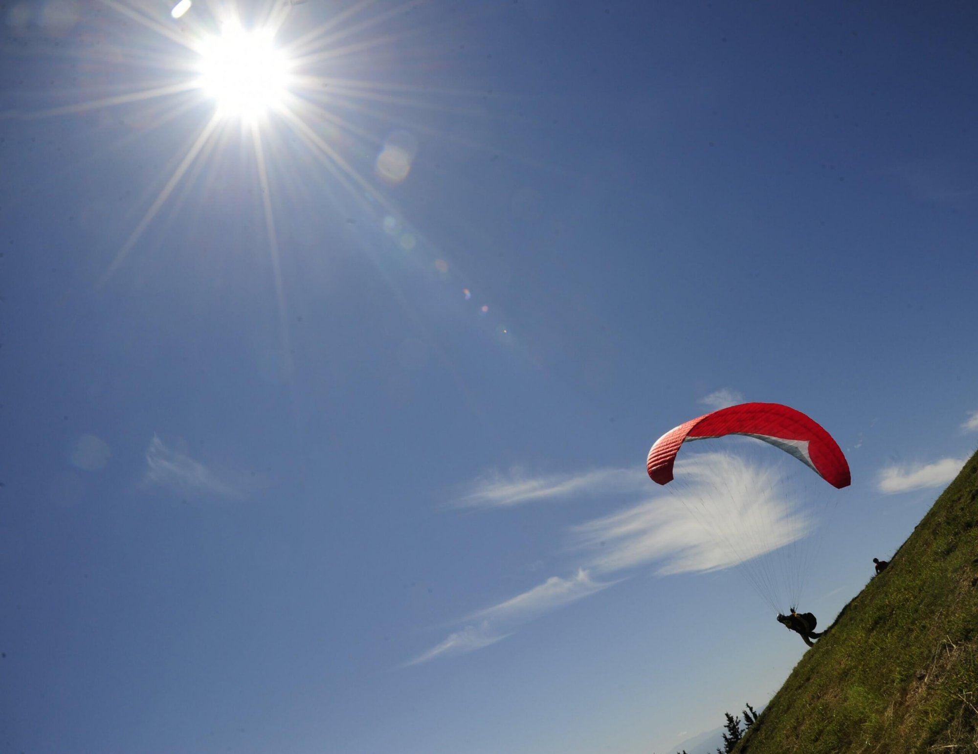 Paragleiter mit rotem Schirm startet von einer Wiese bei Sonnenschein und blauem Himmel.